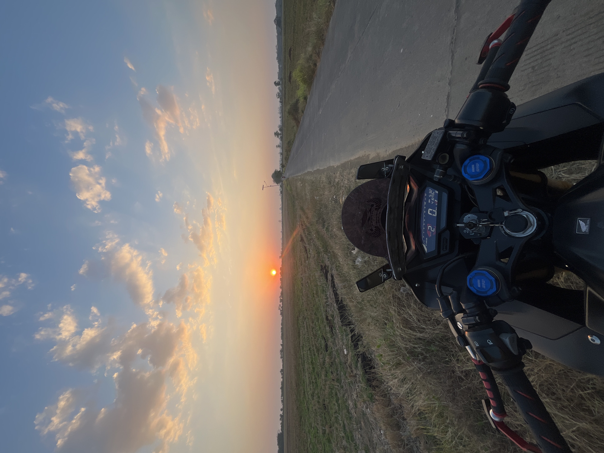 The image depicts a motorcycle parked on the side of a paved road in the city of Pecangaan, Indonesia, during a sunset. The viewpoint is from the rider's perspective, showing the handlebars and dashboard of the motorcycle in the foreground. The road extends into the distance, bordered by dry, grassy fields on both sides. In the background, the sun is setting low on the horizon, casting a warm, orange glow across the sky, which is filled with scattered, wispy clouds. The overall scene conveys a sense of tranquility and the end of the day in a rural Indonesian setting.