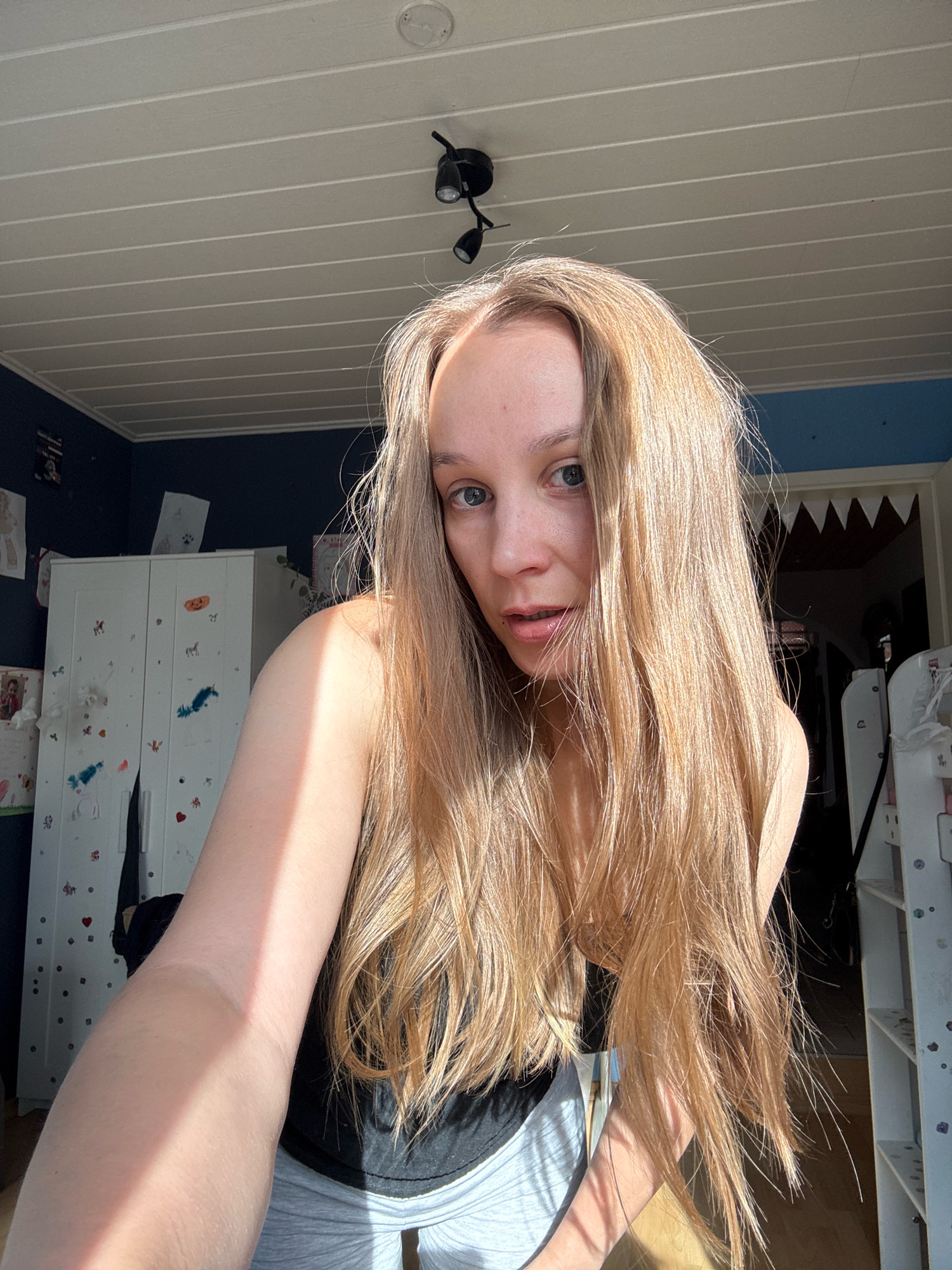 The image shows a young woman with long, blonde hair, wearing a black top and grey sweatpants, taking a selfie in a room with a sloped, wood-paneled ceiling. The room appears to be a child's bedroom, with a white wardrobe decorated with stickers, including a pumpkin, horses, and feathers. The walls are dark blue. There is a ceiling light fixture with multiple spotlights. Sunlight streams into the room, casting a strong shadow across the woman's arm and hair. The overall impression is a casual, indoor setting. There is no visual information to suggest a location in Überherrn, Germany.