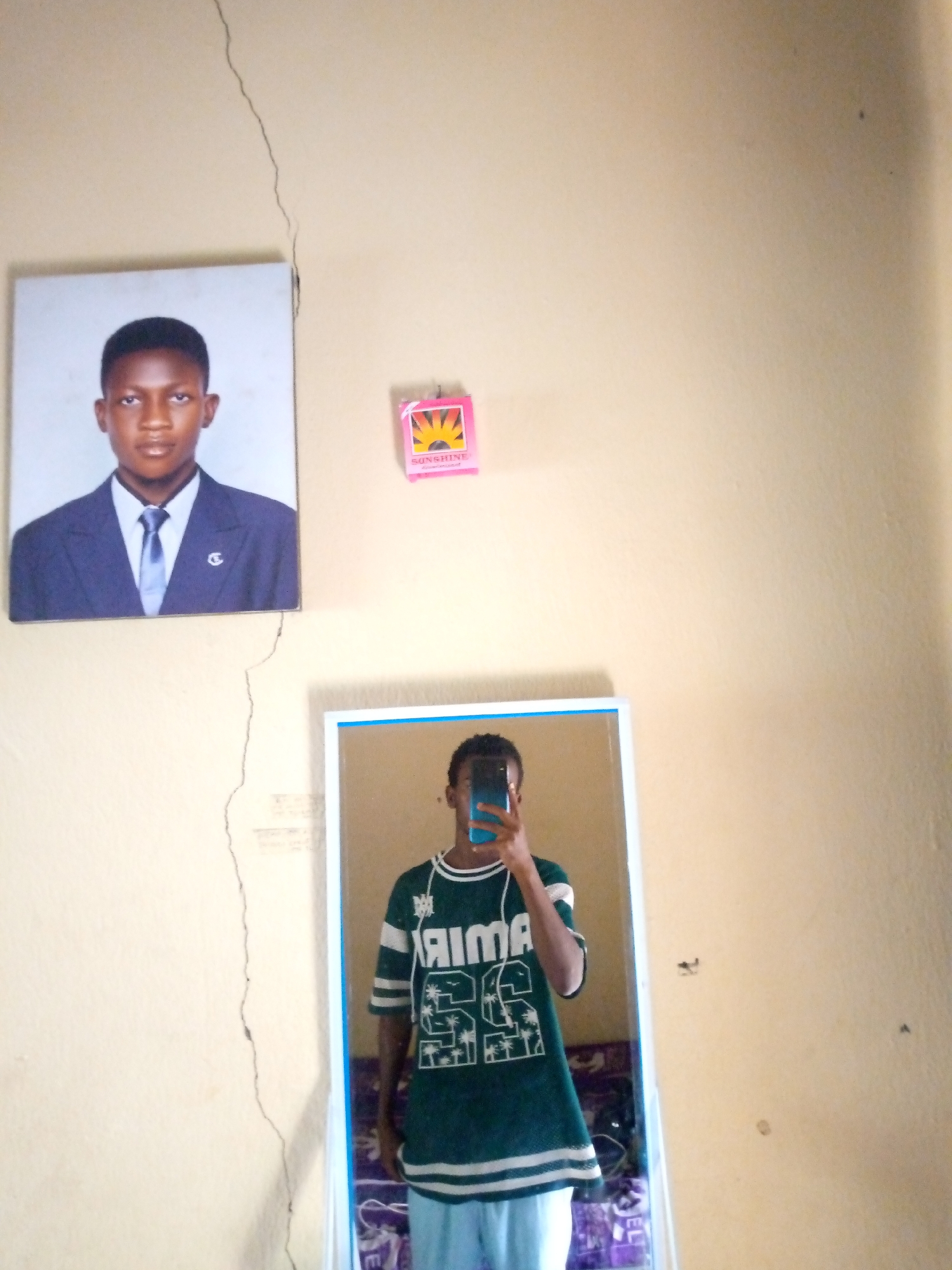 A young Black male is captured in a vertical, medium-shot, indoor photograph. The subject is wearing a dark green t-shirt with white accents and text, light blue pants, and is holding a blue smartphone to take the picture. The background is a plain, light-colored wall with a noticeable crack running vertically down the center. To the left of the crack, a framed portrait of the same young male, dressed in a suit and tie, is hung on the wall. Above the portrait and to the right, a small, pink box with a sun graphic and the word "SUNSHINE" is affixed to the wall. Below the portrait, a full-length mirror reflects the subject, indicating the photograph is a selfie. The environment appears to be a domestic setting, possibly a bedroom in Jalingo, Nigeria, given the context. The lighting is adequate, illuminating the scene clearly.