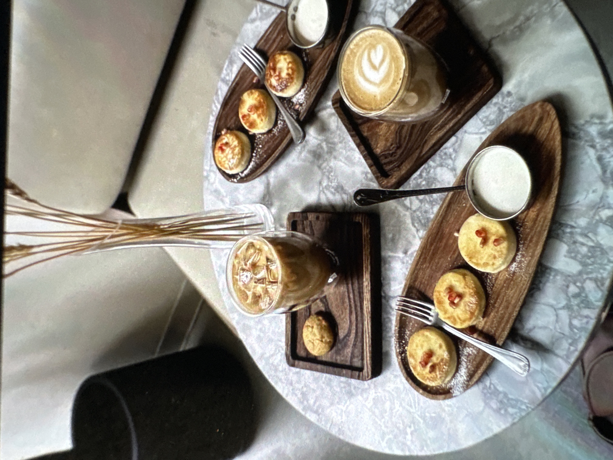 The image depicts a table setting in what appears to be a cafe in Yerevan, Armenia. Two types of small, round pastries are arranged on wooden platters. Some are topped with small red garnishes, possibly pomegranate seeds, and dusted with powdered sugar. Alongside each set of pastries is a small container of white sauce. Silver forks are placed next to each platter. There are also two glasses of coffee. One contains a latte with latte art, while the other is an iced coffee. The wooden platters holding the drinks are distinct in size and shape from those holding the pastries. The table itself has a marbled surface. A vase containing wheat stalks is visible in the background. Part of a seat and a dark cylinder-shaped object (possibly a wastebasket or other furniture) are also in the background.