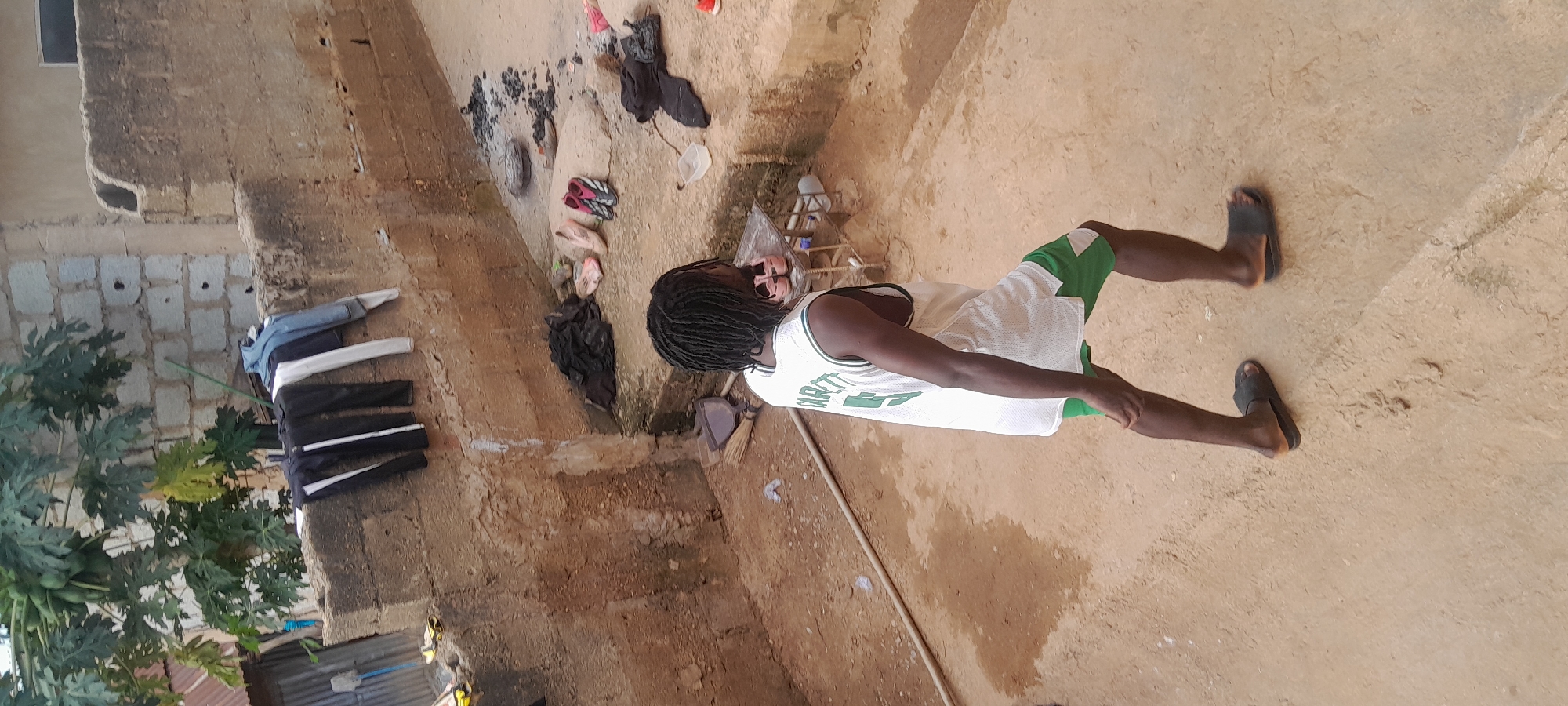 An outdoor scene in Abuja, Nigeria, features a person walking away from the camera in a courtyard. The person is wearing a white tank top with green trim and the name "GARRETT" and the number "5" on it, and green shorts. They are also wearing dark-colored sandals. To the left, several pieces of clothing, including pants and a jacket, are hanging on a stone wall. A papaya tree with green fruit is visible behind the wall. In the foreground and mid-ground, the ground is composed of packed dirt and sand, with some areas appearing damp. Various objects are scattered around, including a broom, a dustpan, a few pairs of shoes, and some dark-colored fabric. In the background, a rough, unfinished brick wall and a section of corrugated metal roofing are visible. The overall impression is of a functional outdoor living space.