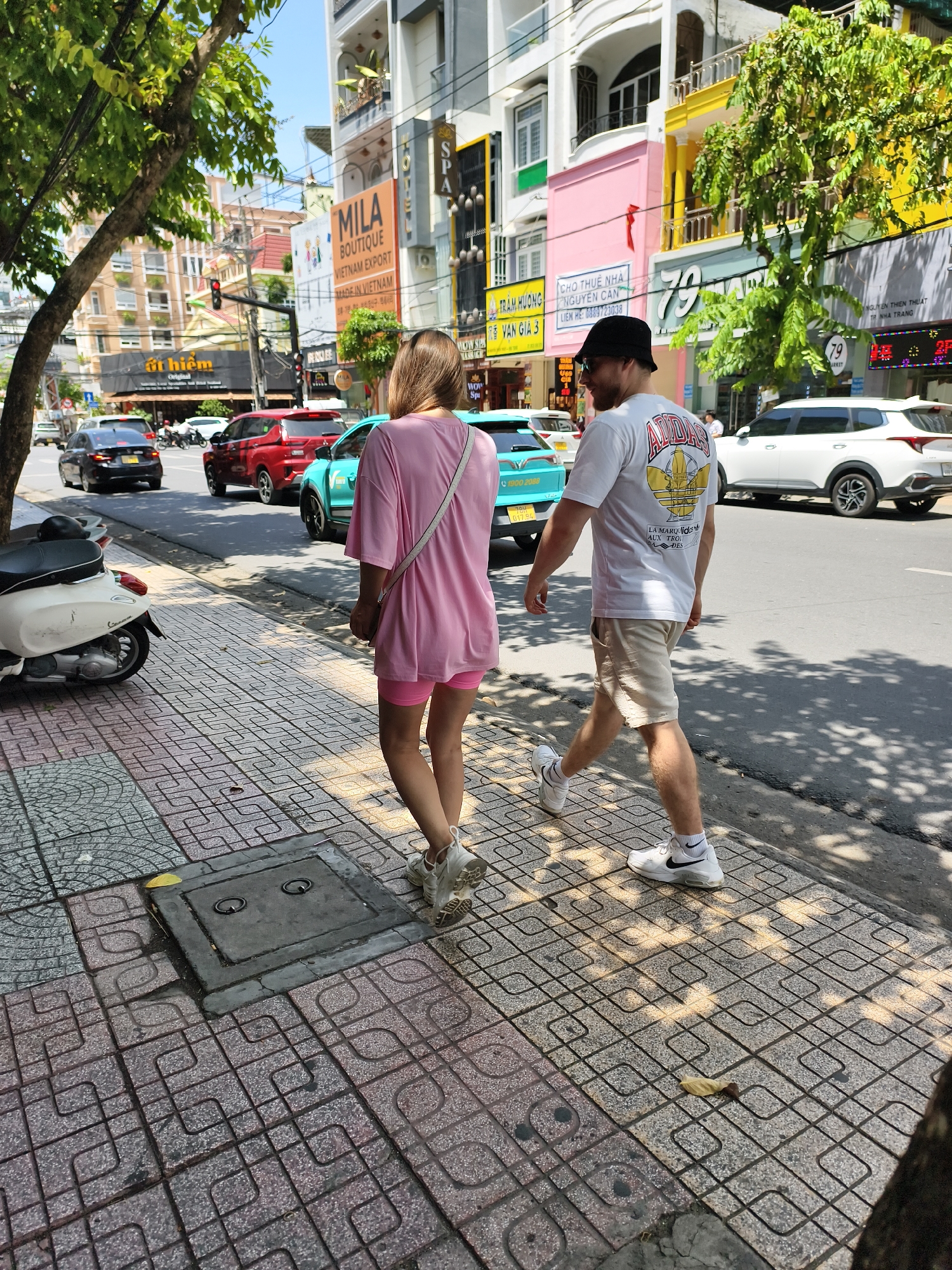The image shows a street scene in Nha Trang Ward, Vietnam. A man and a woman are walking on a patterned sidewalk along a busy street with cars, trucks, and motorbikes. The woman is wearing a pink outfit with a white shoulder bag. The man is wearing a white Adidas shirt, khaki shorts, and a black hat. In the background are buildings with signs for "MILA BOUTIQUE", "CHO THUÊ NHÀ", "TRẦM HƯƠNG NGUYỄN CAN", and "79 MARKET". A white scooter is parked near the sidewalk. The weather appears sunny, with shadows from trees cast on the sidewalk.