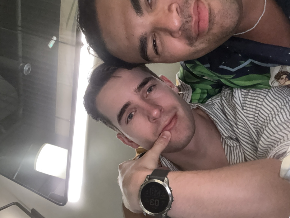 Two adult males are depicted in a close-up selfie, likely taken indoors within a vehicle. The man on the left has light skin, dark short hair, and blue eyes; he wears a light-colored vertically striped shirt and a silver-toned smartwatch with a black band on his left wrist. His right index finger is positioned against his lower lip. The man on the right has a darker complexion, dark hair, and dark eyes; only the right half of his face is clearly visible, and he wears a dark patterned shirt, possibly with green and white tropical foliage, and a thin white beaded necklace. Both individuals are looking towards the camera. The background shows a reflective overhead panel, revealing a rectangular light source and a circular fixture, consistent with the interior design and panoramic window of a Dubai Metro train. The scene is located within a Dubai Metro train in Dubai, United Arab Emirates.