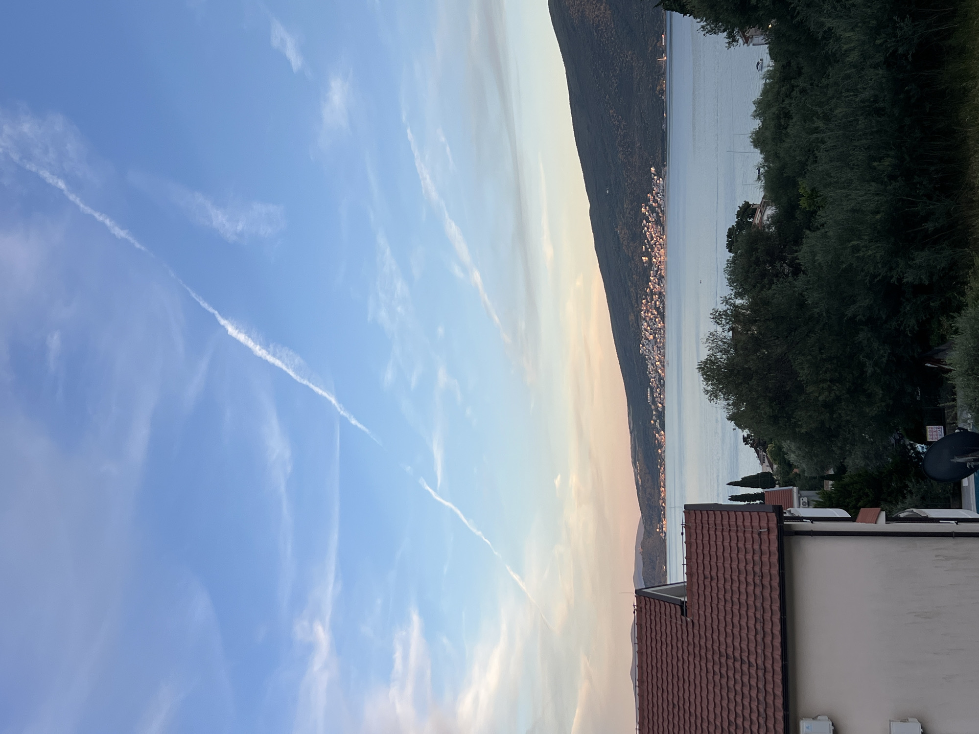 This image depicts a serene coastal scene in Baošići, Montenegro. In the foreground, the roof of a building with red tiles is visible on the left. Beyond the building, lush green trees dominate the middle ground. Further in the distance, a body of calm water stretches out, with a town nestled along the coastline of a verdant mountain that rises from the opposite shore. The sky above is a soft blue, streaked with wispy clouds and a distinct contrail from an aircraft. The lighting suggests it is either early morning or late afternoon, casting a gentle glow over the landscape. No people are clearly visible in the frame.