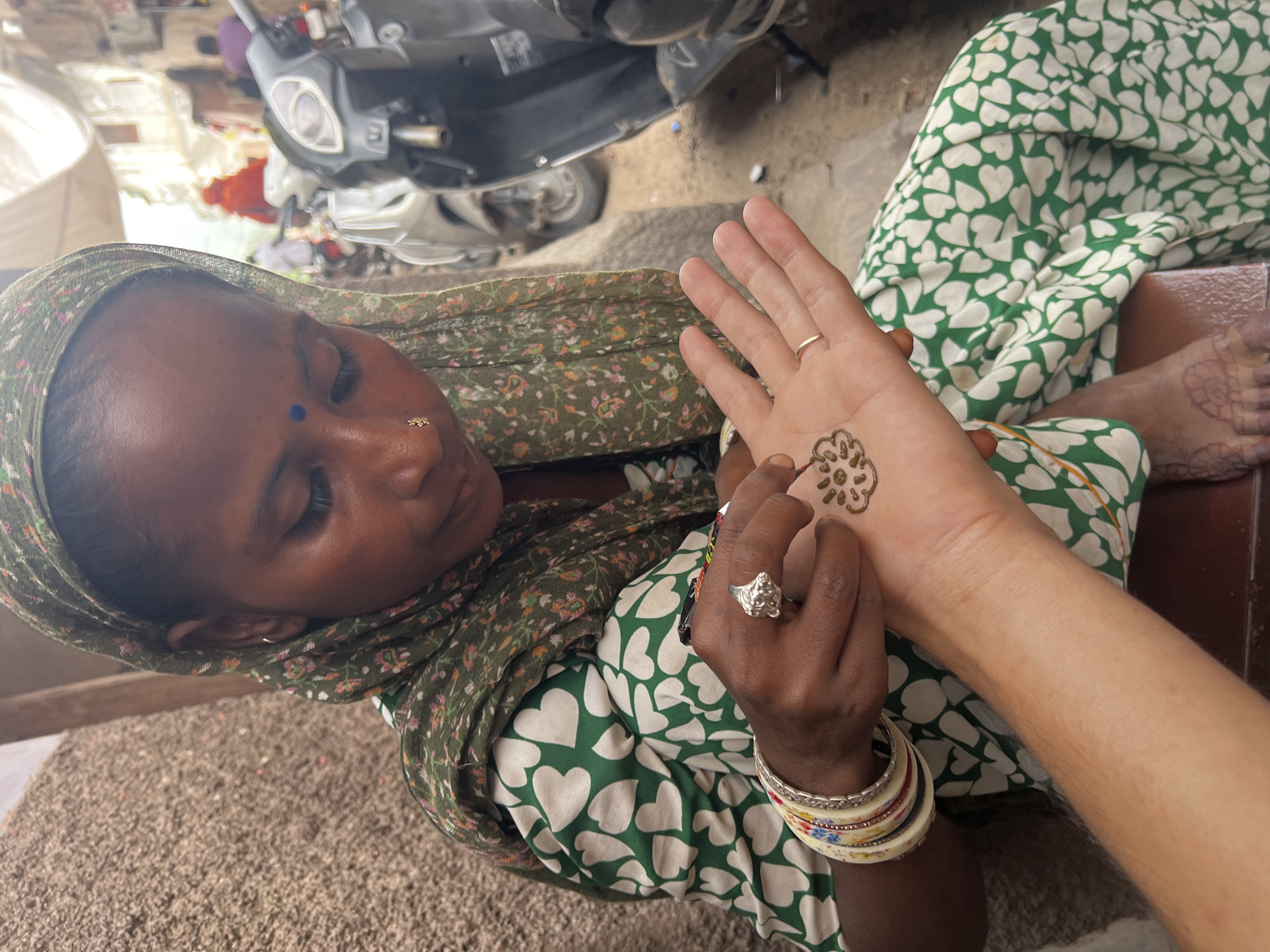 A woman in Pushkar, India, applies henna to a person's palm. The woman, wearing a green patterned headscarf and dress, concentrates on her task. The person's palm, with a thin gold ring on one finger, is being decorated with an intricate henna design. In the background, a busy street scene unfolds with motorcycles and people. The environment appears to be an outdoor market or street stall.
