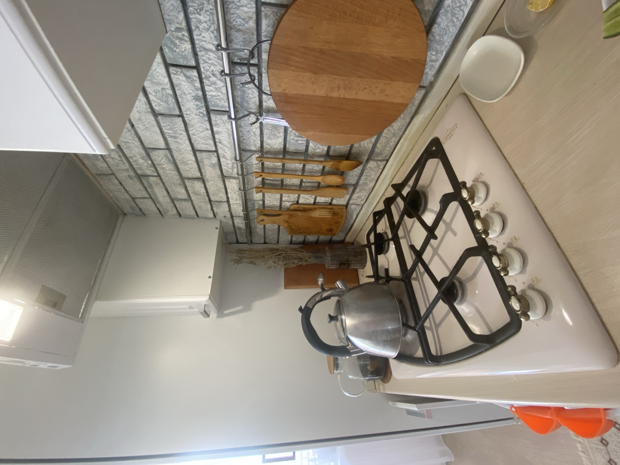 The image shows a close-up view of a kitchen counter and stovetop. The stovetop is white and has four burners with black grates. A silver teapot is on one of the burners. To the left of the stovetop is a glass teapot with a wooden lid, containing tea. Behind the stovetop, the backsplash is made of light-colored, textured brick. A metal rack is mounted on the backsplash, holding several wooden utensils and a round wooden cutting board. The counter is a light wood tone. In the foreground, to the right of the stovetop, a small white oval dish and a clear dish with a slice of lemon and cucumber are visible. To the left of the stovetop, near the edge of the counter, two orange plastic scoops are stacked. A white range hood is visible above the stovetop, and a white cabinet is to its left. The overall impression is a clean and organized kitchen space.