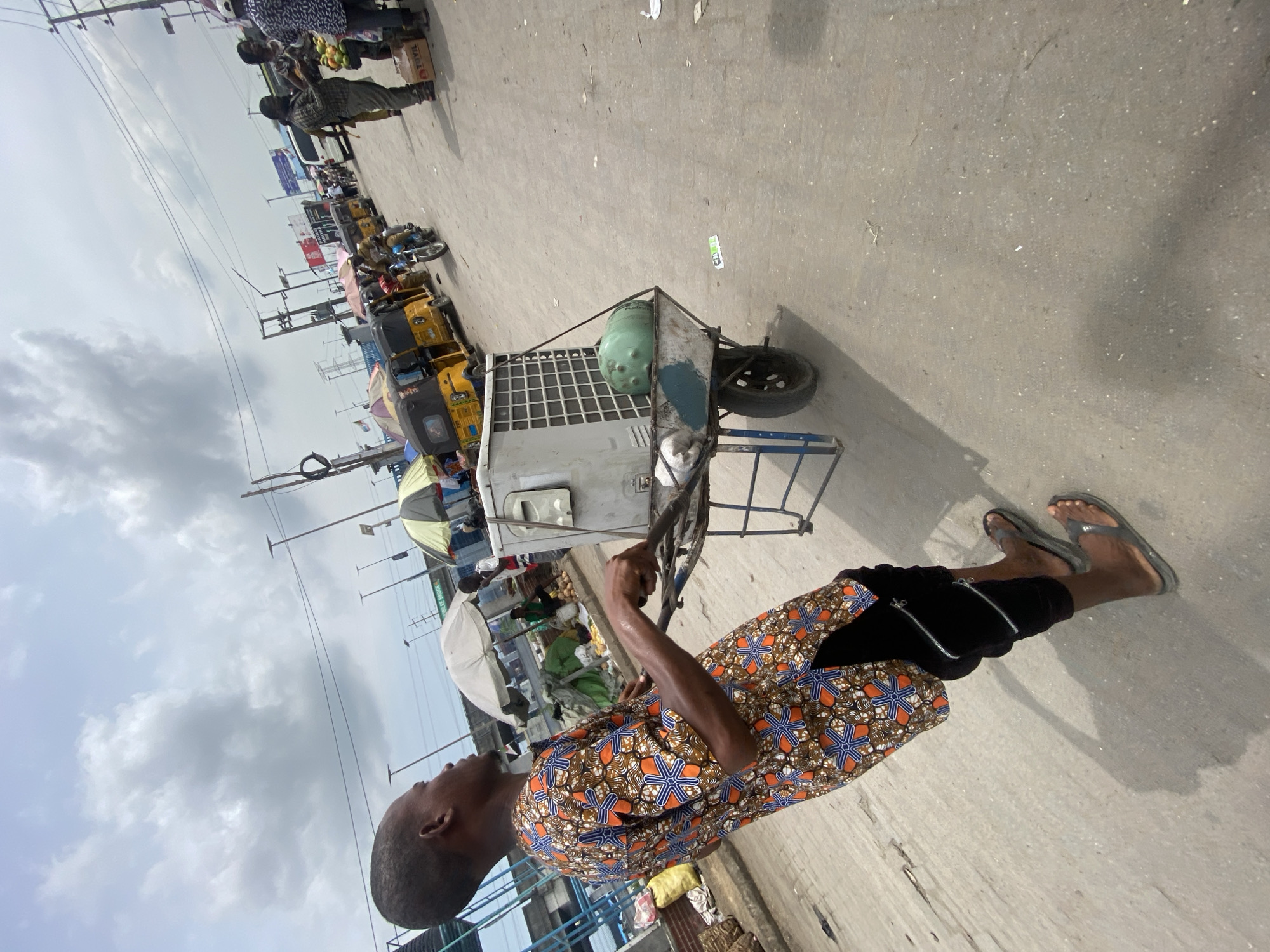 The image captures a lively outdoor street or market scene in Lagos, Nigeria, under an overcast sky. In the foreground, a man of African descent is seen pushing or guiding a two-wheeled handcart. He wears a vibrant, patterned short-sleeved shirt featuring orange, blue, and brown geometric designs, black pants with zippered sides, and dark flip-flops. His gaze is directed upwards and to his left, with a focused expression.
The handcart, a utilitarian metal frame structure, carries a large, light-colored rectangular object, possibly a cooler or an appliance with a gridded top, and a light green, barrel-shaped container behind it. The cart appears well-used.
The background portrays a bustling urban environment. Several other people are visible, including vendors attending market stalls. One vendor is seen with a display of fruits. Multiple yellow tricycles, commonly known as "Keke Napep" in Nigeria, are parked or moving along the street, alongside a blue motorcycle, suggesting active local transportation. Market stalls, some shaded by white or striped umbrellas, line the thoroughfare. Overhead, numerous power lines crisscross the grey, cloudy sky, supported by utility poles. The paved street surface appears worn and scattered with small debris. Visible text includes the word "TOTAL" on a brown box in the background, and part of a sign reading "FREE MUSIC." The overall scene depicts a typical day of commerce and movement in a Nigerian city.