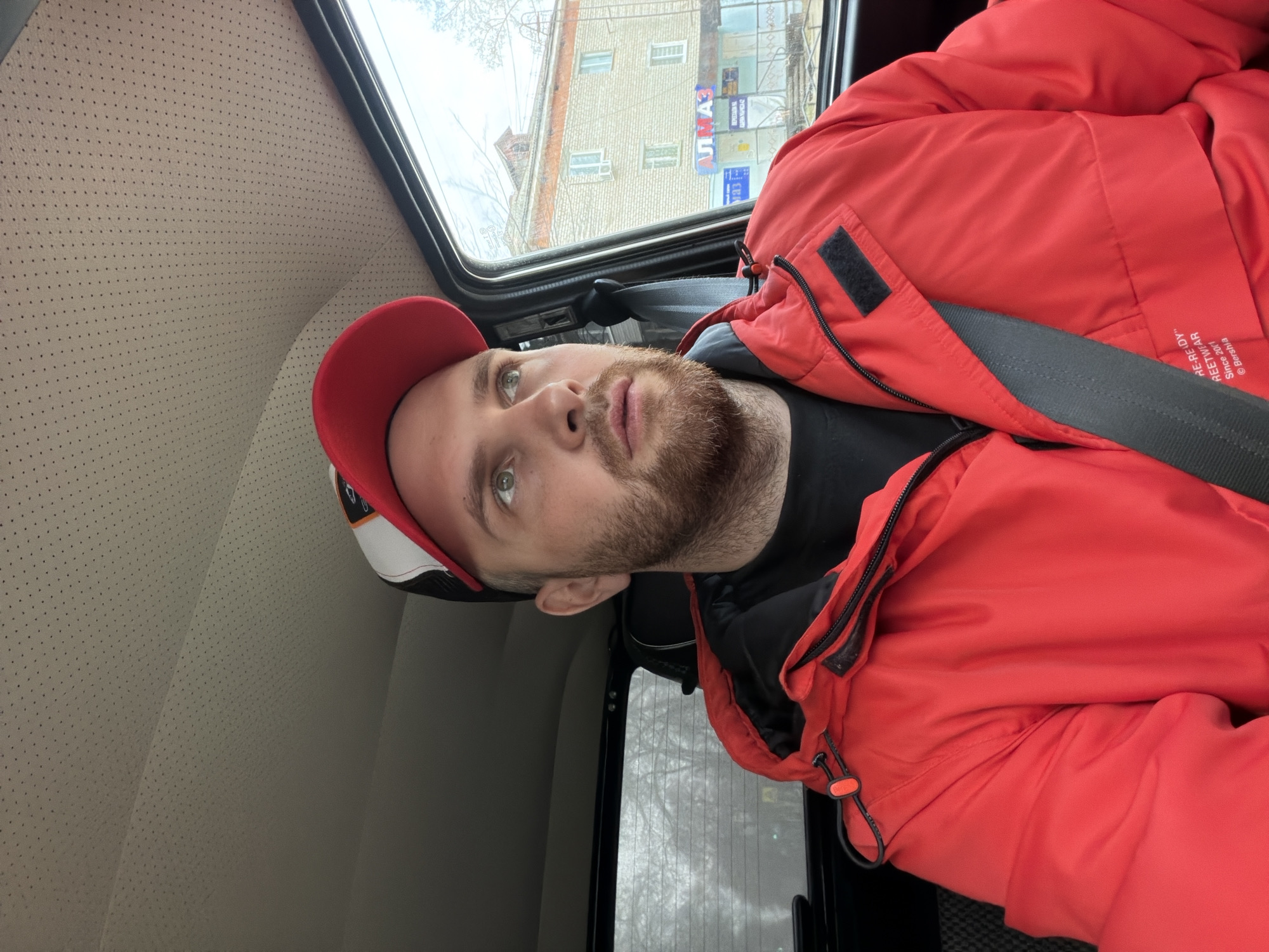 A man with a beard and a red baseball cap is depicted in a portrait-style shot, taken inside a vehicle, likely in Chaikovsky, Russia. He is wearing a bright red, possibly padded, jacket over a black t-shirt, and a seatbelt crosses his chest. The man is looking upwards and slightly to his left with a neutral, observant expression.
The interior of the vehicle features a light beige, perforated headliner or side panel, and a dark window frame. Through the window, an urban street scene is visible. The exterior environment includes a light-colored brick building with multiple windows, suggestive of residential or commercial architecture common in Russian cities. Bare trees indicate late autumn, winter, or early spring, and the diffused light suggests an overcast daytime.
Visible text on the man's red jacket, near the shoulder, reads "RE-RE-DY", "REETWEAR", "since 2011", and "© Bershka", identifying the brand. Outside the window, on a street sign, the Cyrillic word "АЛМАЗ" (ALMAZ) is clearly visible, though mirrored, further confirming the Russian location context. The man appears to be a passenger, observing the surroundings while traveling.
