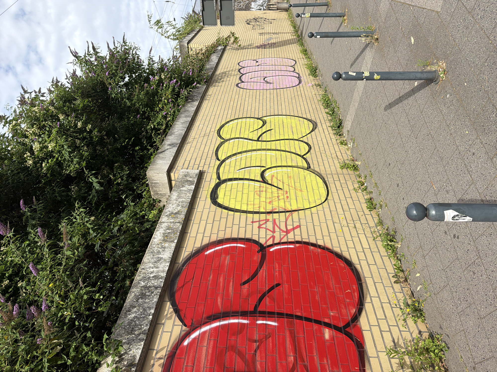 The image shows a brick wall covered in graffiti in Witten, Germany. The graffiti is in red, yellow, and pink bubble letters. The wall is located next to a sidewalk, which is paved with gray interlocking concrete pavers. There are several bollards along the sidewalk, and some vegetation is growing along the base of the wall. In the background, there are trees and a cloudy sky.