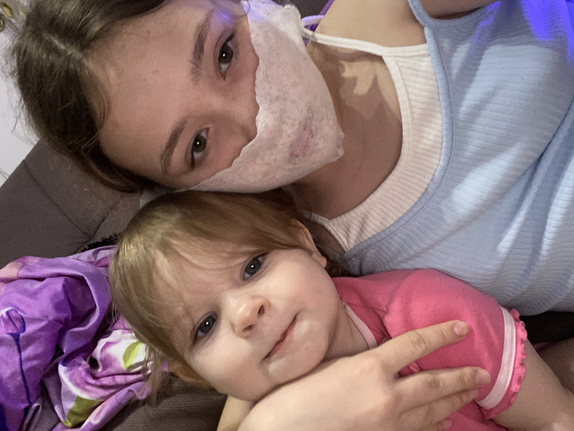 Two individuals are depicted indoors. An older female with brown hair is wearing a light blue top and a white facial covering that is partially torn. A younger female with blonde hair is wearing a pink top with white trim. They are lying together on what appears to be a couch, which has a patterned purple blanket nearby. One person's arm is visible, seemingly cradling the child. The location is assumed to be indoors in Unknown, Unknown.