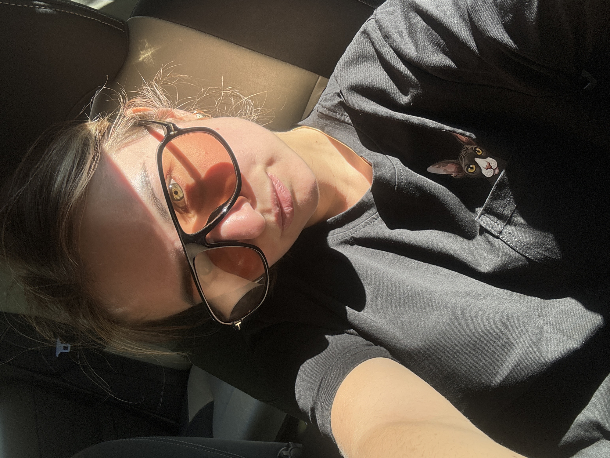 In Villard-Saint-Sauveur, France, a young woman with light brown hair is taking a selfie inside a vehicle. She is wearing large, dark-rimmed sunglasses with orange lenses and a dark gray t-shirt that has a printed image of a cat peeking out of the breast pocket. The sun is shining brightly, casting strong shadows across her face and clothing, and illuminating the interior of the car. The background shows dark seats and a hint of a light-colored seatbelt clip.