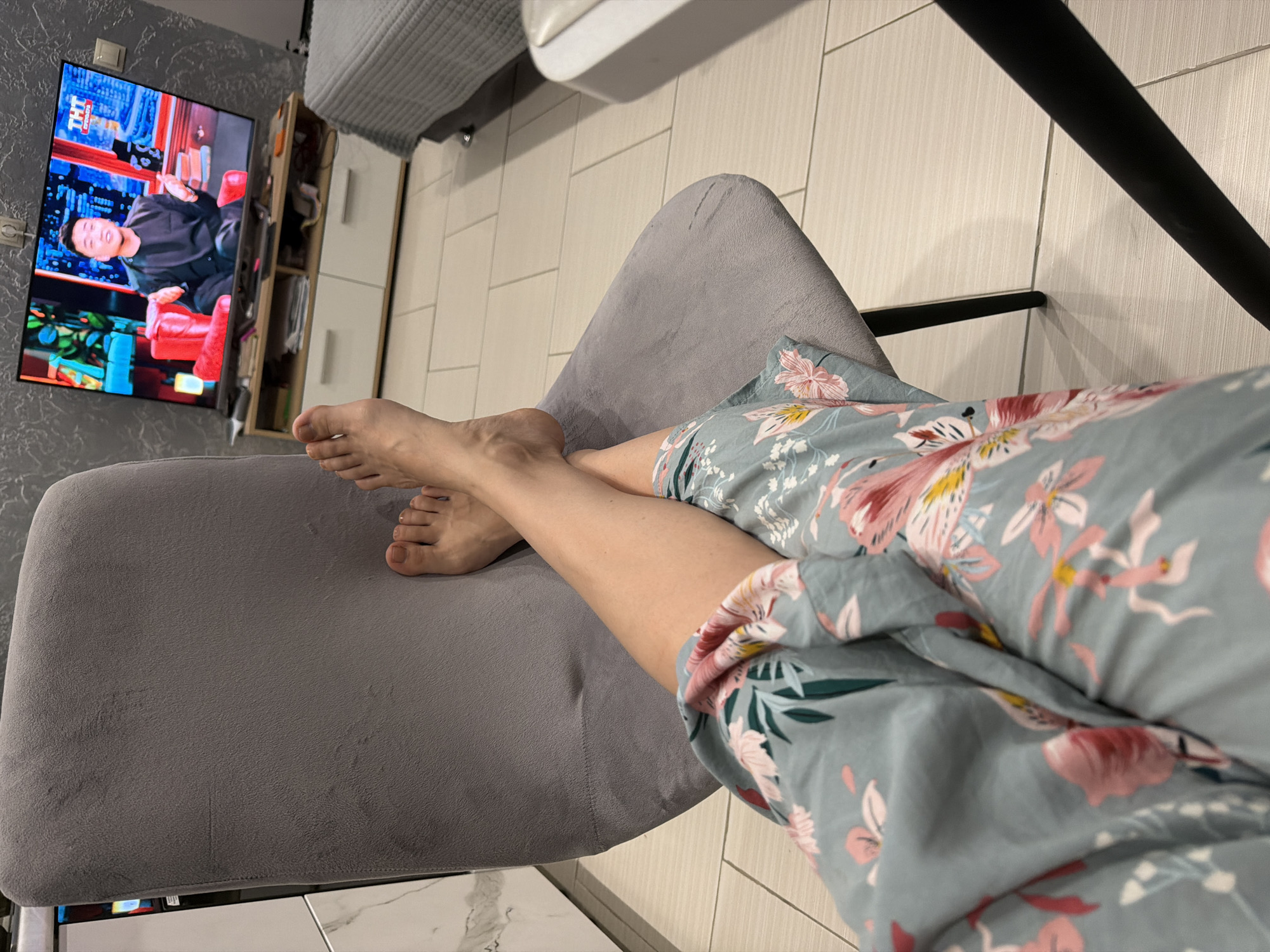 A view from an elevated perspective shows a person relaxing on a gray chair indoors, presumably at home, watching television. The individual is wearing a light blue floral-patterned robe or garment. Their bare feet rest on the chair seat. The television, mounted on the wall, displays a man sitting in a red chair, seemingly from a show or broadcast bearing a "TNT" logo, suggesting the Russian television channel. The apartment's interior design includes light-colored tile flooring and white cabinets. The location may be in Moscow, Russia, based on the TV channel logo.