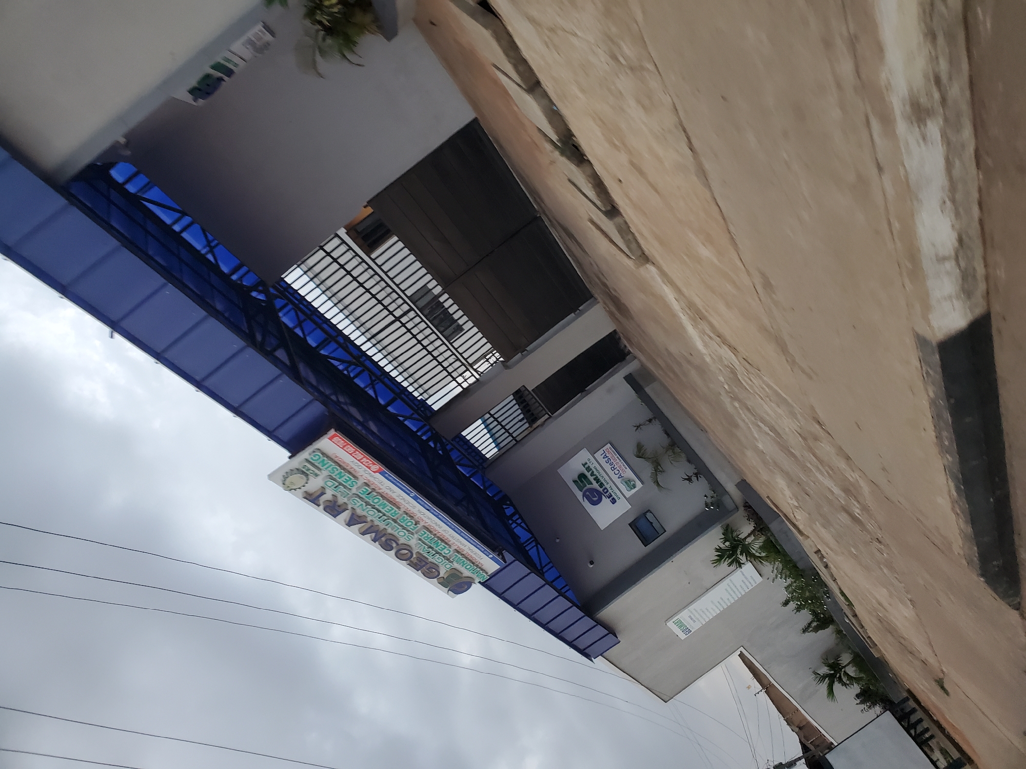 The image shows the exterior of the National Space Research and Development Agency (NASRDA) in Bukuru, Nigeria. The building is modern, with a light gray facade and a blue, corrugated plastic roof extending over a walkway. Several signs are visible. One large sign above the walkway reads "GEOSMART DIGITAL SOLUTIONS LTD" with a smaller inscription below that is partially obscured. Another sign on the building's facade also displays "GEOSMART DIGITAL SOLUTIONS LTD" and further below, "ACRESAL". The building has large, dark metal gates leading into the property. The foreground consists of a paved area with visible cracks and some concrete slabs. Small, manicured plants and palm trees are planted in beds around the building. The sky is overcast.