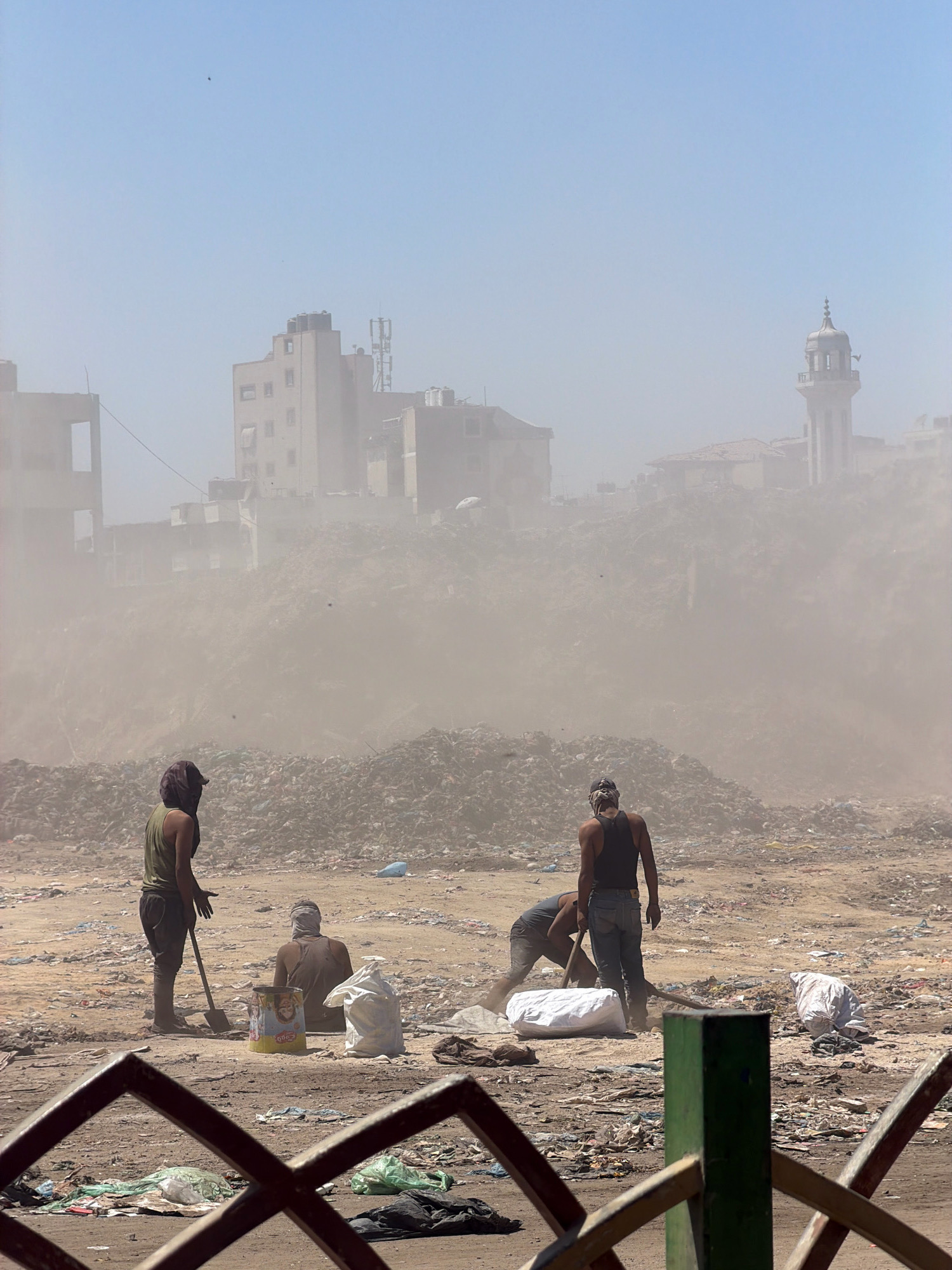 In Gaza City, Palestinian Territories, a group of men are engaged in clearing debris in a dust-filled environment. In the foreground, four men are visible. One man, wearing a green tank top and dark pants, stands with a shovel in hand, appearing to direct or supervise. To his left, another man is seated, his face obscured by a head covering, next to a colorful cylindrical container and a large white sack. Further to the right, two other men are working, one kneeling and the other standing. They are surrounded by piles of rubble and refuse, with scattered trash and debris covering the ground. In the background, damaged buildings and a minaret stand against a hazy sky, suggesting the aftermath of destruction or ongoing demolition. The air is thick with dust, indicating a disturbed or ongoing construction/demolition site.