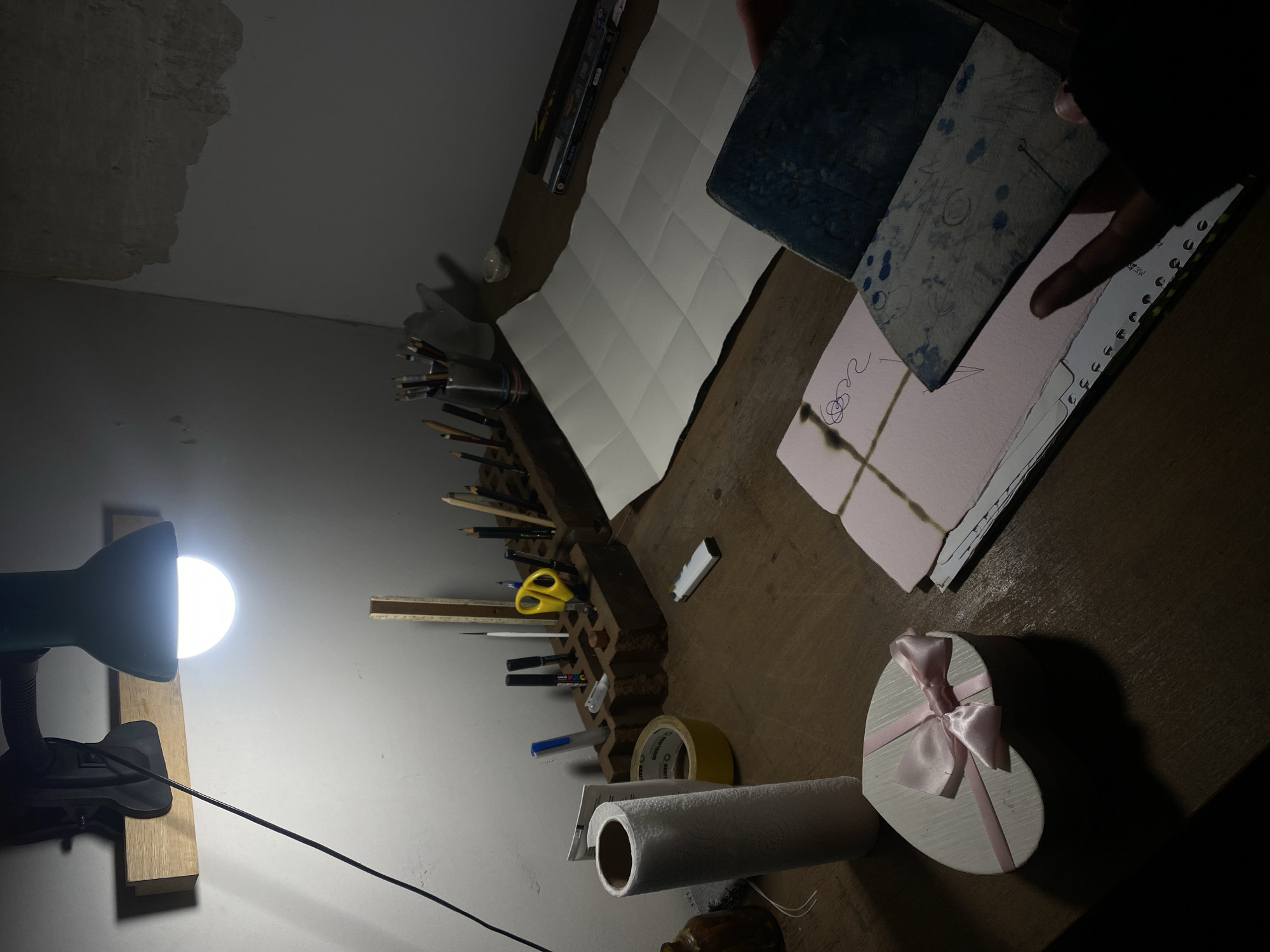 The scene appears to be a workspace with a desk lamp illuminating the area. The workspace contains various art supplies, including pencils, pens, scissors, a ruler, tape, and paper. There is also a box with a pink ribbon. A hand is holding a printing block over a pink piece of paper on the desk. The environment seems to be indoors, but there is no information available in the image that suggests this scene is located in Moscow, Russia.