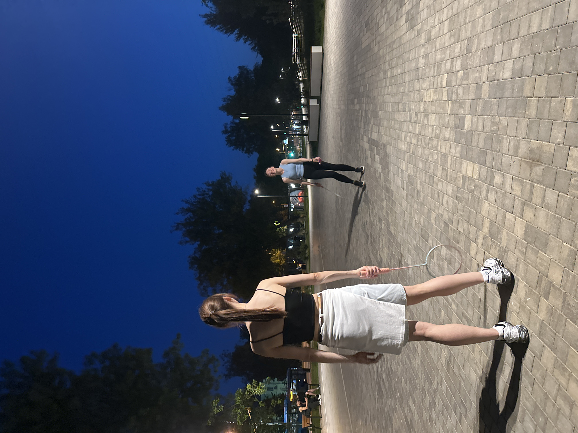 Two young women are playing badminton in a paved outdoor area at dusk. The woman in the foreground is facing away from the camera, holding a badminton racket. She is wearing a black tank top, a white wrap skirt, and athletic shoes. The woman in the background is facing the camera and appears to be laughing. She is wearing a light blue tank top and black leggings. The scene is lit by streetlights, and trees and buildings are visible in the background. The location is identified as a park or public square in Krasnogorsk, Russia.
