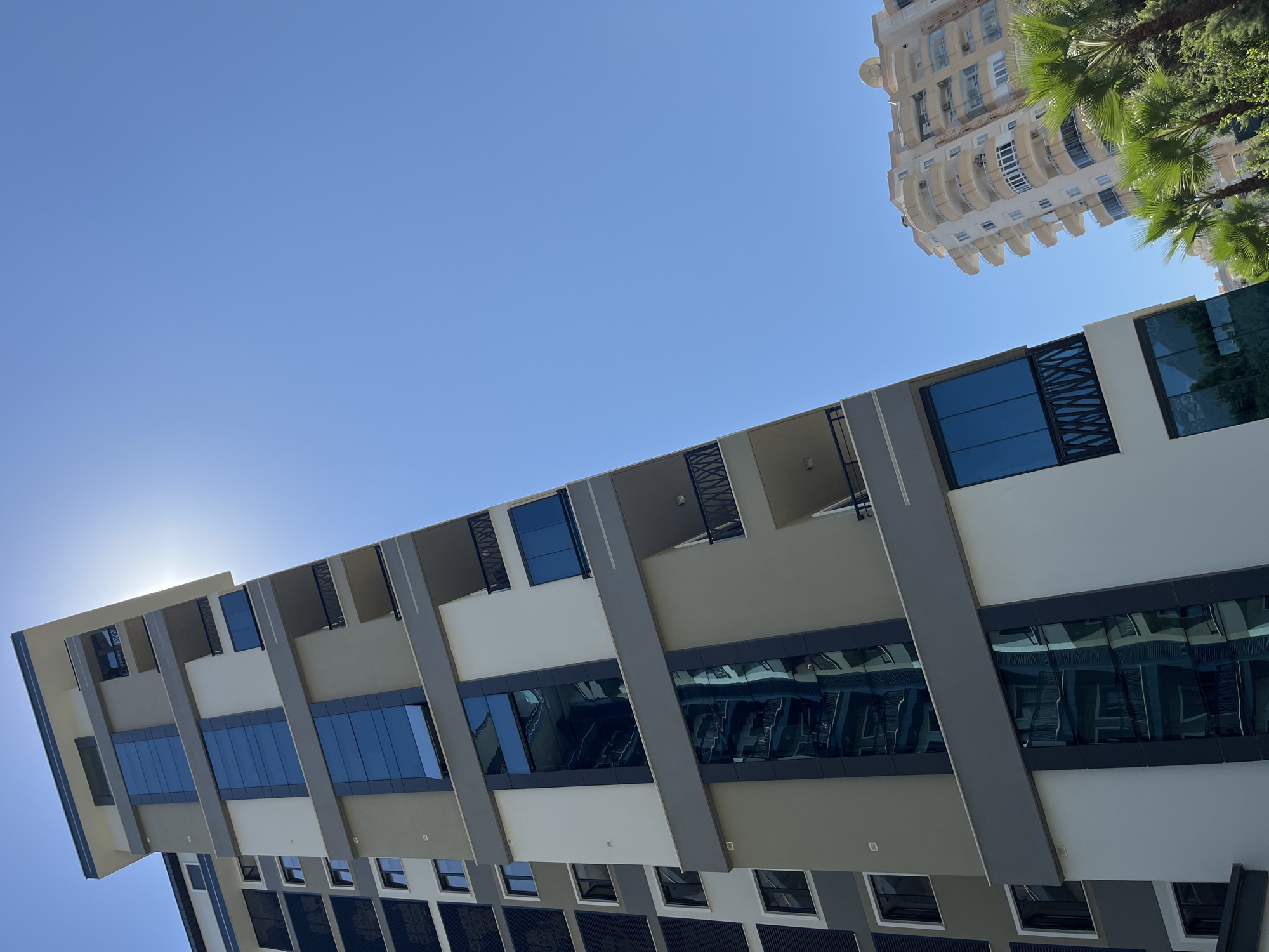 This image depicts a modern residential building in Alanya, Turkey, with a clear blue sky overhead. The building is tall and features a contemporary design with a mix of light gray, white, and dark gray sections. Large windows, some with a reflective blue tint, are prominent elements of the facade. Balconies with black railings are visible on several floors.
To the right of the main building, another lighter-colored, multi-story building with rounded balconies is partially visible. In the foreground and to the lower right, lush green palm trees and other foliage add a tropical feel to the environment. The sun is high in the sky, casting a bright light and creating a slight glare on the top portion of the main building. The angle of the photograph is from a low perspective, looking upwards at the buildings. There are no people visible in the image. The overall scene suggests a sunny day in a developed urban area with vegetation.