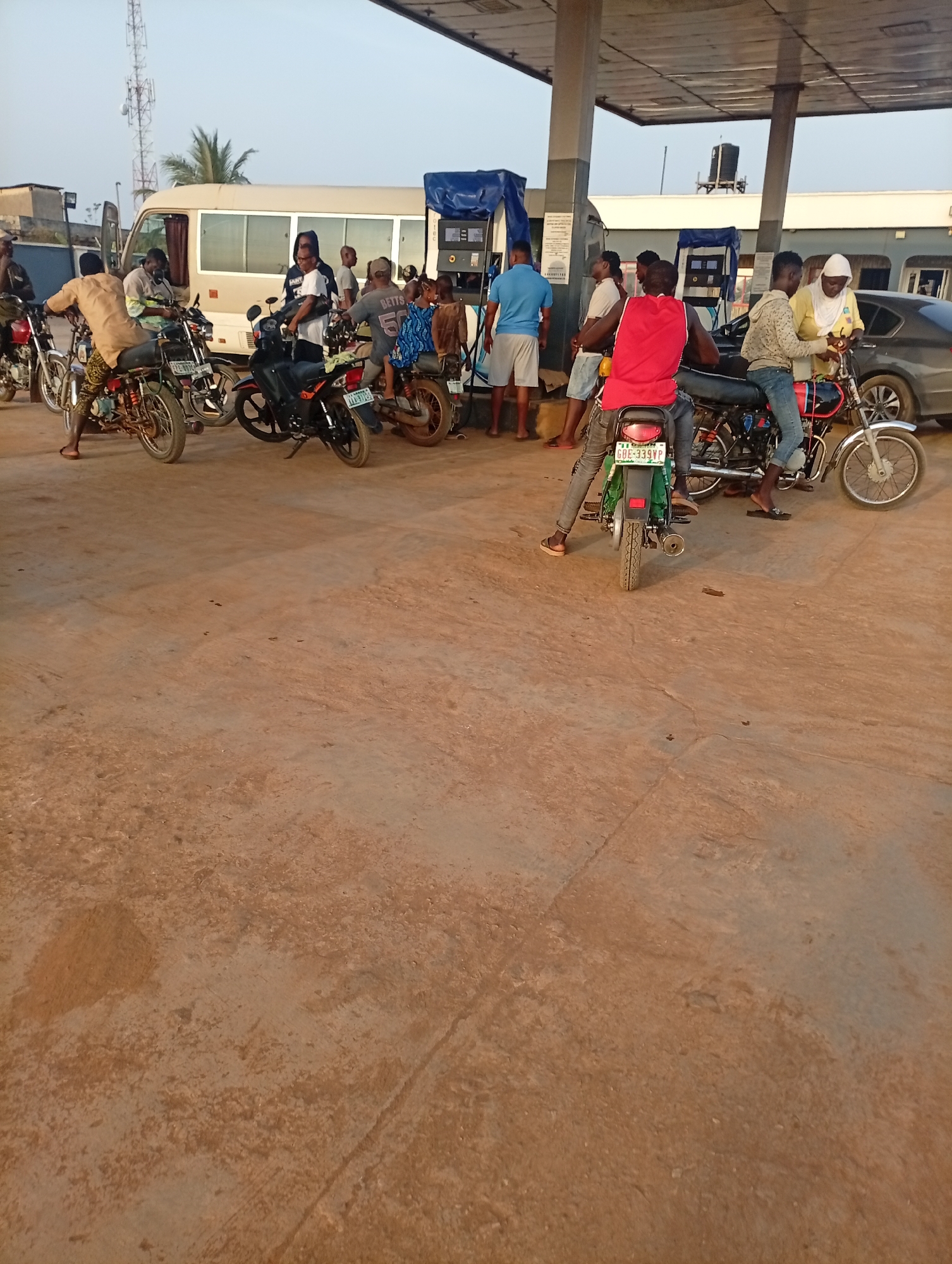 The image depicts a scene at a petrol station in Ikorodu, Nigeria. Several individuals are gathered around fuel pumps, primarily refueling motorcycles. A white van is parked nearby, and a dark-colored car is also present. The ground is a light brown, possibly concrete or dirt. The overall scene suggests a bustling environment where people are engaged in refueling activities at the petrol station.