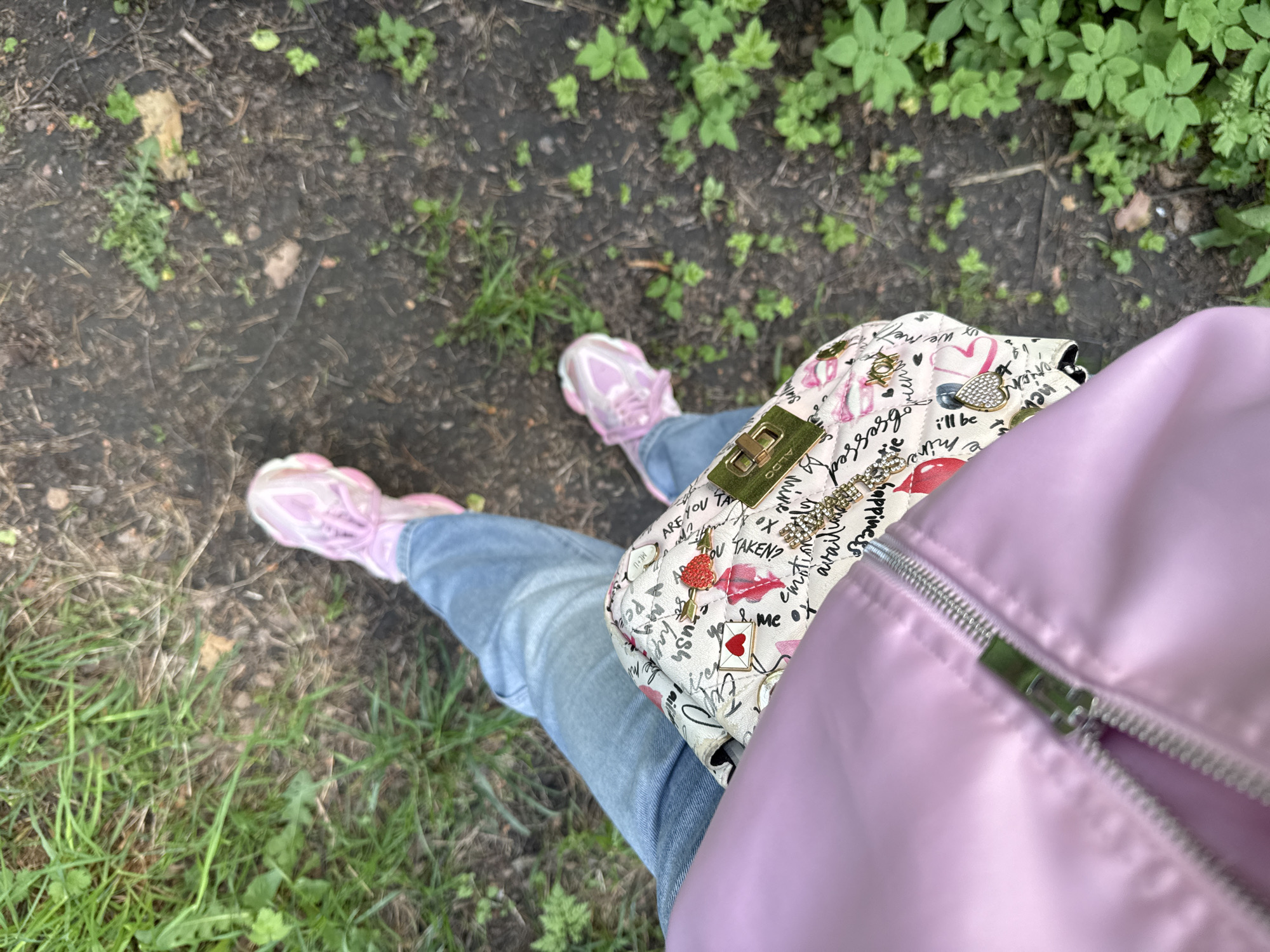 The image depicts a person standing on a path with patches of grass and dirt. The individual is wearing light blue jeans, pink sneakers, and a lavender-colored jacket. They are carrying a white purse with various graphic designs. The scene appears to be outdoors, potentially in a park or garden. The text on the bag includes words or phrases such as "obsessed," "mine," and "happiness awaits." The location is not specified.