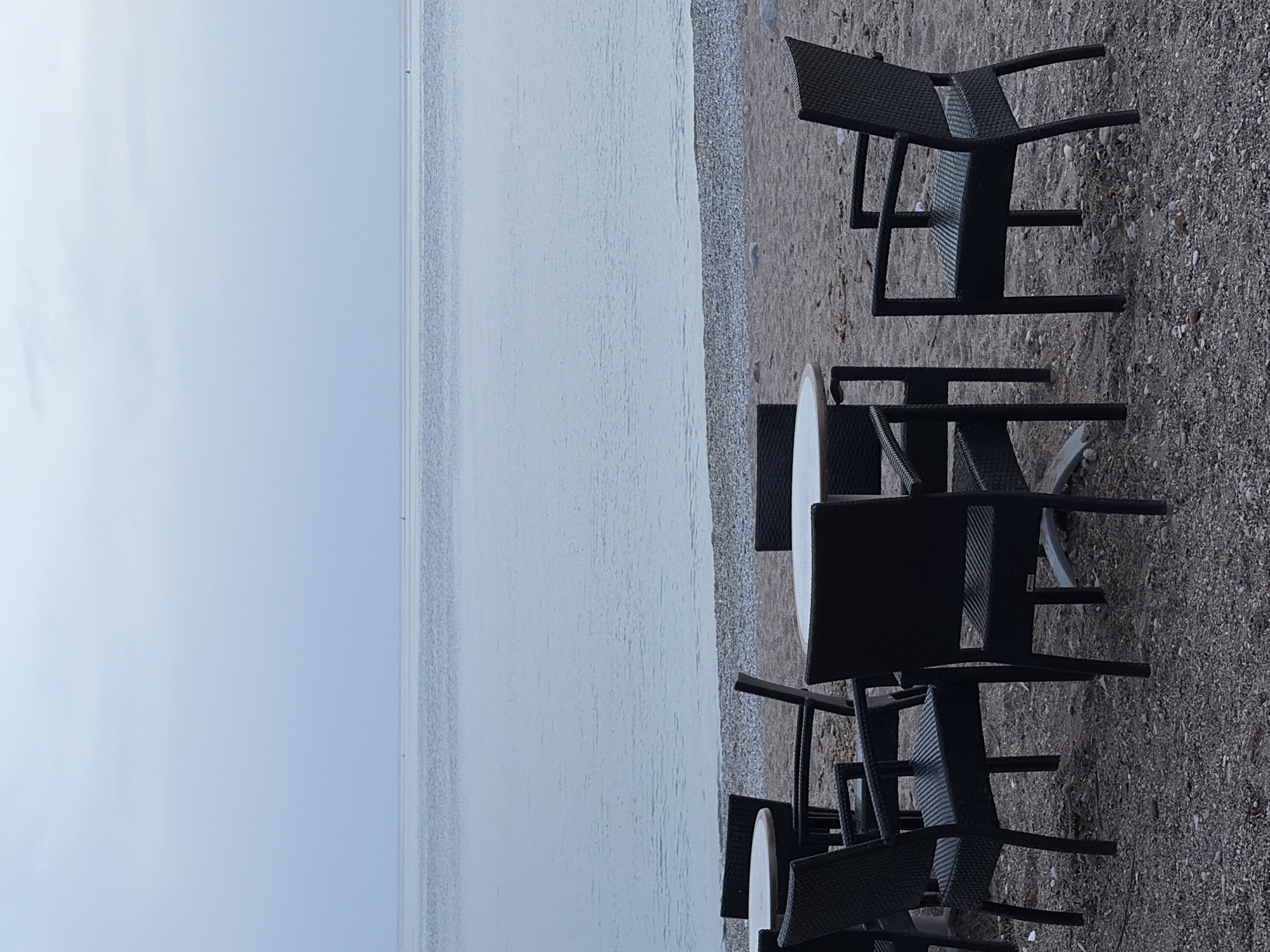 The scene depicts outdoor dining furniture – tables and chairs – on a gravel beach in Budva, Montenegro. The furniture is of a dark, possibly black, woven material. The beach extends to a calm body of water, likely the Adriatic Sea, which fades into a gray sky. The image is taken from a low angle, emphasizing the furniture and its proximity to the water. There are no people visible in the image.