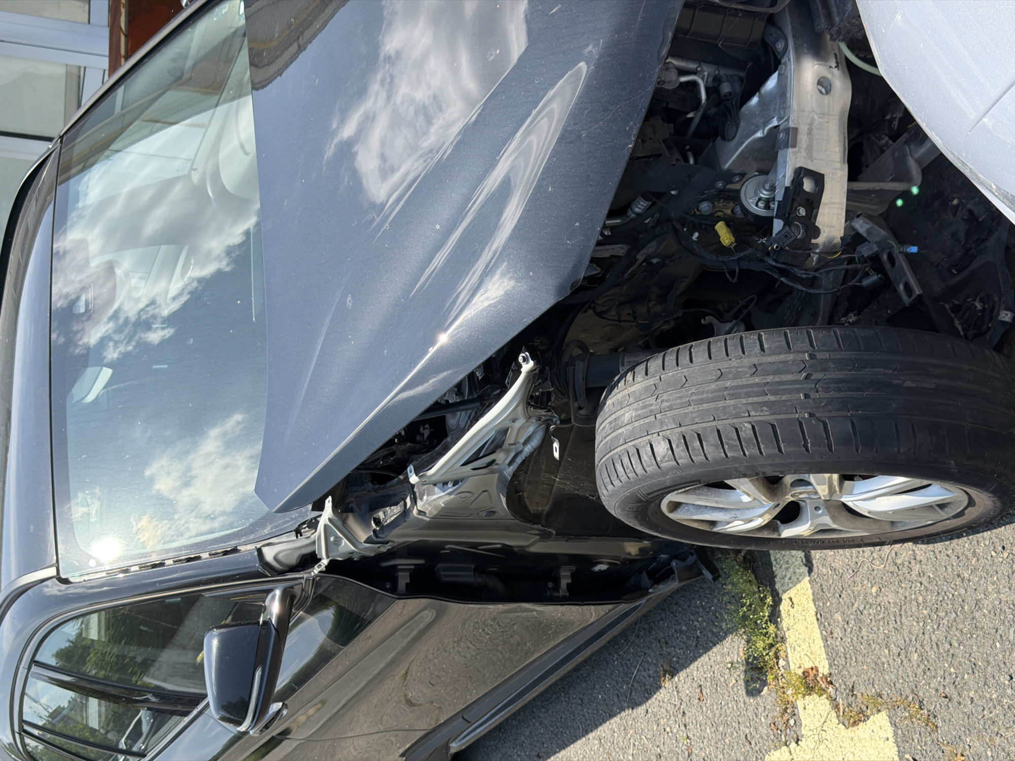 This media file captures a close-up, angled view of a severely damaged dark grey or black car, likely the aftermath of an accident. The car is situated outdoors on what appears to be asphalt pavement with a faded yellow line, indicating a roadside or parking area. The time of day is clearly daytime, with bright sunlight creating reflections of clouds and surrounding structures on the car's body panels and windows.
The most prominent feature is the extensive damage to the front right side of the vehicle. The hood is crumpled, misaligned, and partially open, exposing the engine bay and various internal components, including wires, hoses, and a bent, silvery chassis member. The front right wheel and tire are visibly detached from their original mounting position, standing independently near the exposed engine area. The tire exhibits typical road wear, and the silver alloy wheel is visible.
Reflections on the side windows clearly show fluffy white clouds in a bright sky, as well as vague outlines of what might be buildings or other vehicles. The car's exterior appears dusty. A small portion of a white vehicle is visible on the far right edge of the frame, suggesting another vehicle might be involved or parked nearby. The location context provided is Burgas, Bulgaria. There is no clearly legible text visible on the car or in its immediate surroundings. The overall impression is one of significant vehicular destruction.