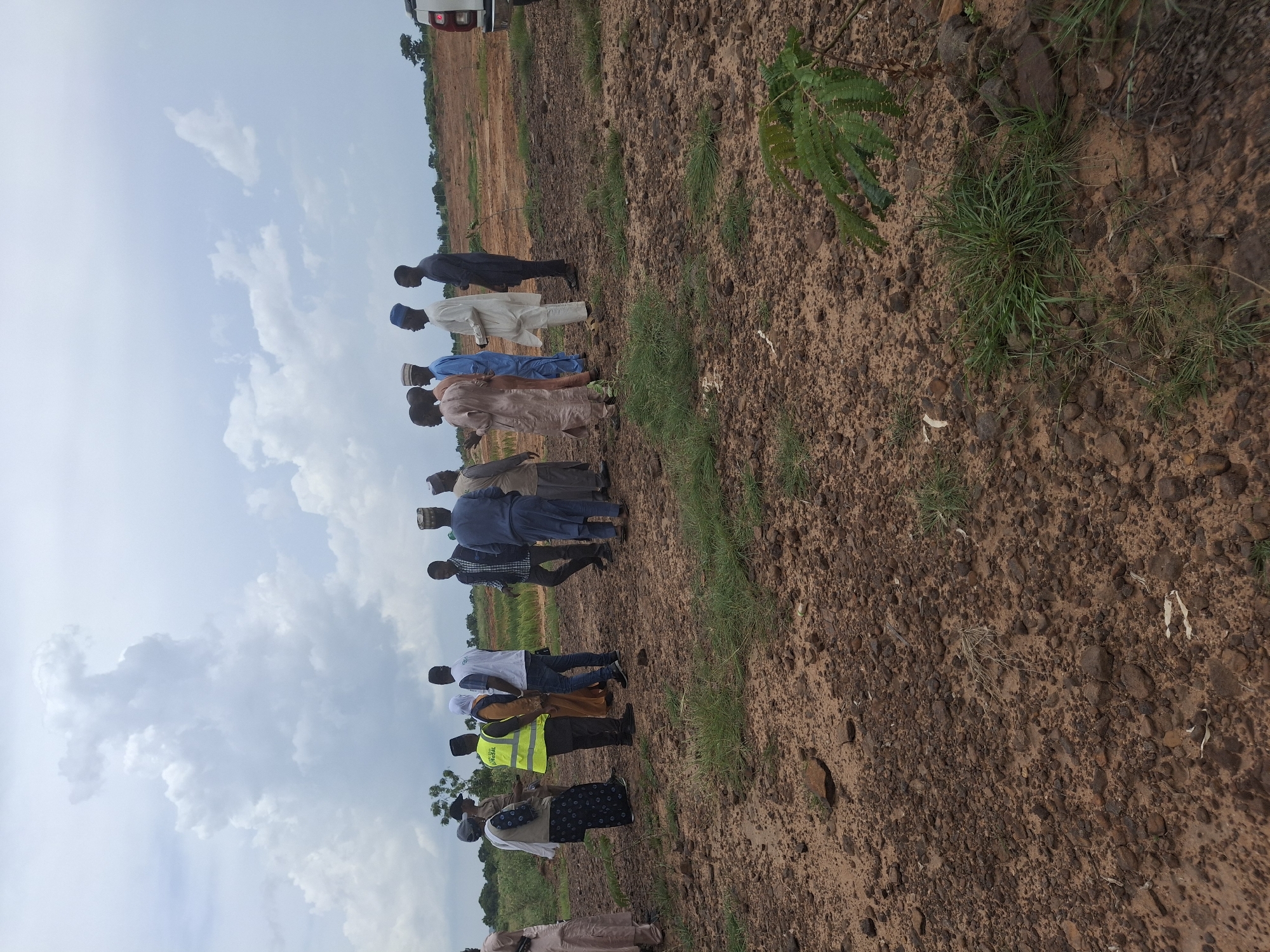 A group of approximately ten people, dressed in a mix of traditional African attire and Western clothing, are gathered in an open, undeveloped field in Danaje, Nigeria. The ground is predominantly brown soil mixed with stones and sparse patches of grass. In the background, there is evidence of construction or land preparation, with cleared earth and distant trees. The sky is overcast with dramatic, dark clouds. Some individuals appear to be in conversation, while others are observing the surroundings. One person is wearing a bright yellow safety vest. A white vehicle is partially visible on the right side of the frame. The overall impression is a site visit or meeting taking place in a rural or semi-rural area.