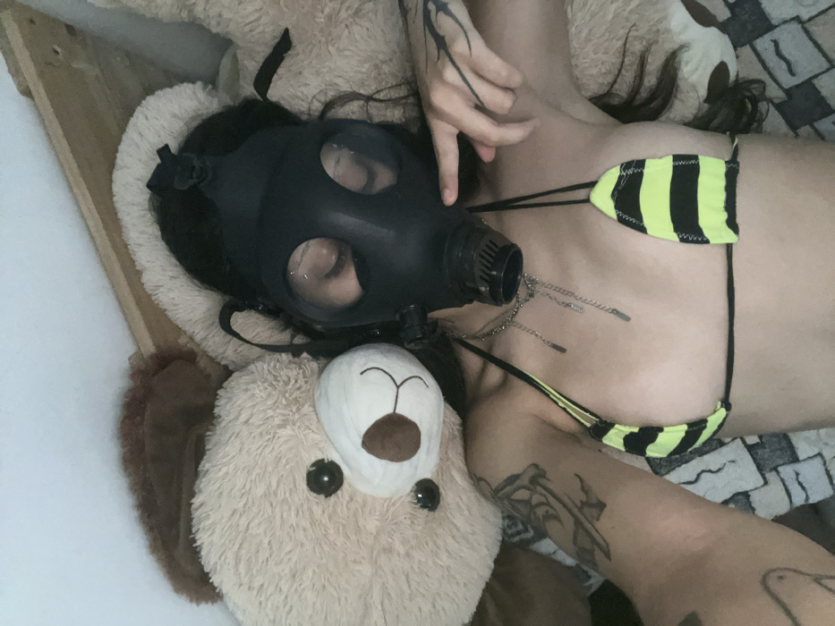 A person lies down wearing a black gas mask, with their face visible through the lenses. A large, light brown teddy bear with a white snout and black eyes rests beside the person's head. The person is wearing a black and neon yellow striped bikini top. Their arms and upper chest feature tattoos. The setting includes a textured blanket or sheet.