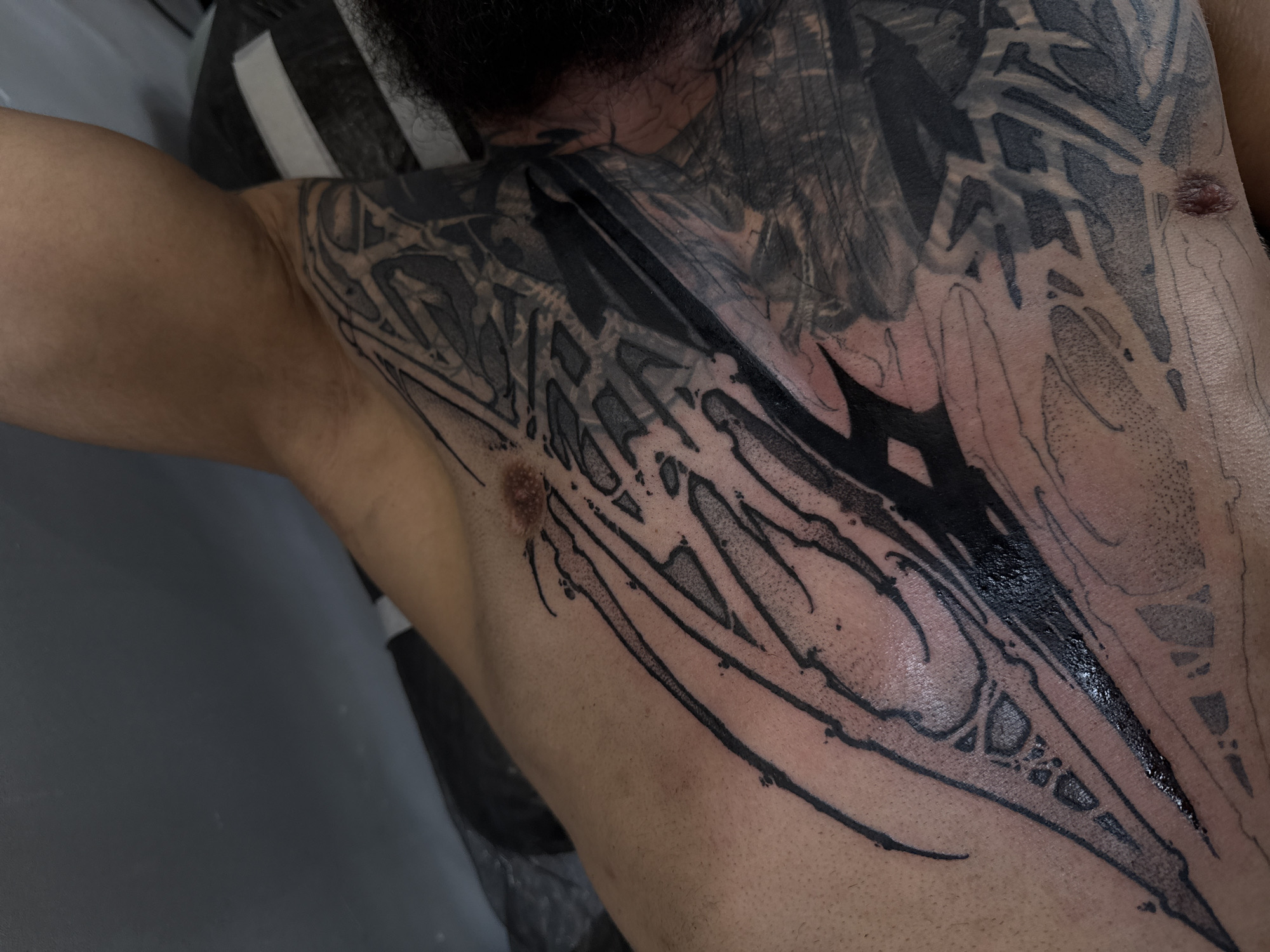The image shows a person's chest and shoulder with a large black and grey tattoo. The tattoo design is intricate and appears to be a combination of tribal and geometric patterns. The person's skin is fair, and a small amount of dark hair is visible near the neck and shoulder area. The image also captures parts of a grey background. The location is not specified, so Moscow, Russia is not indicated.