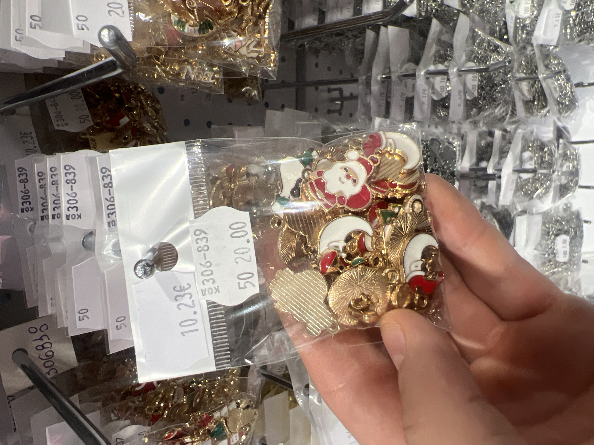 Here is a forensic description of the image:
The image appears to have been taken inside a store, possibly in Burgas, Bulgaria, based on the price tag format (€ symbol). The main subject is a hand holding a small, clear plastic bag filled with Christmas-themed, gold-colored charms and pendants, likely for jewelry making or crafts. The charms include images of Santa Claus, moons, and other seasonal shapes. The bag has a price tag attached, indicating a cost of 10.23€ and a reference number "306-839," with "50 20.00" and the number "50" displayed on other attached tags.
The store setting is evident by the presence of multiple similar bags hanging on metal racks in the background. These bags contain an assortment of different charms, some in gold tones and others in silver. The racks are arranged to display the merchandise for customers. The overall lighting is bright, typical of a retail environment.
