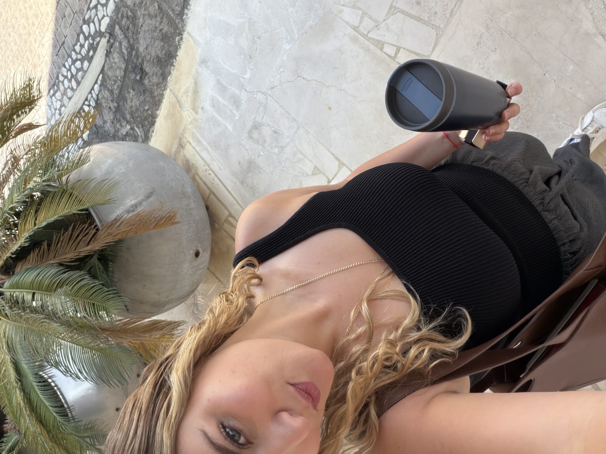 A selfie taken outdoors captures a woman from a high-angle, looking down at the camera. She has blonde, curly hair, blue eyes, and is wearing a black ribbed tank top with a gold chain necklace. Her right hand, adorned with a thin red bracelet, holds a black thermos with a grey lid, which has the word "THERMOS" partially visible. She is also wearing dark pleated pants or a skirt and white sneakers. A brown tote bag hangs from her left shoulder, with a glimpse into its contents.
The setting appears to be a sunny outdoor area in San Bernardino Tlaxcalancingo, Mexico. The ground beneath her is paved with a mix of light-colored, irregularly shaped flagstones and some darker, possibly cobblestone, sections. To her left, a large concrete planter contains vibrant green and dried palm fronds, suggesting a tropical or subtropical climate. The lighting indicates it is daytime with clear weather. Her expression is neutral, engaging directly with the camera.