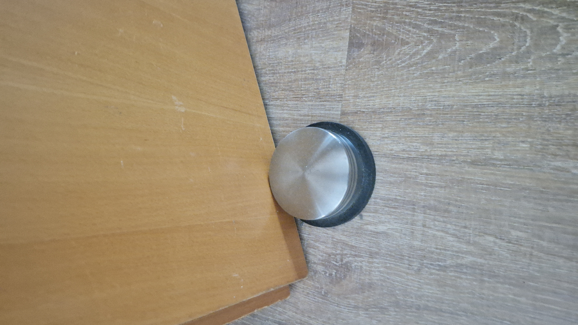 The image shows a stainless steel doorstop with a black rubber base resting on a wood-patterned floor. The doorstop is positioned near the bottom edge of a wooden door, which appears to be a light brown or oak color. The door is partially open, angled away from the viewer. The floor is made of laminate or vinyl flooring with a visible wood grain texture. The lighting is neutral, suggesting indoor ambient light. There are no people or any other discernible objects in the scene. The location is not specified within the image itself, and therefore its presence in Baden-Baden, Germany cannot be confirmed from the visual information provided.
