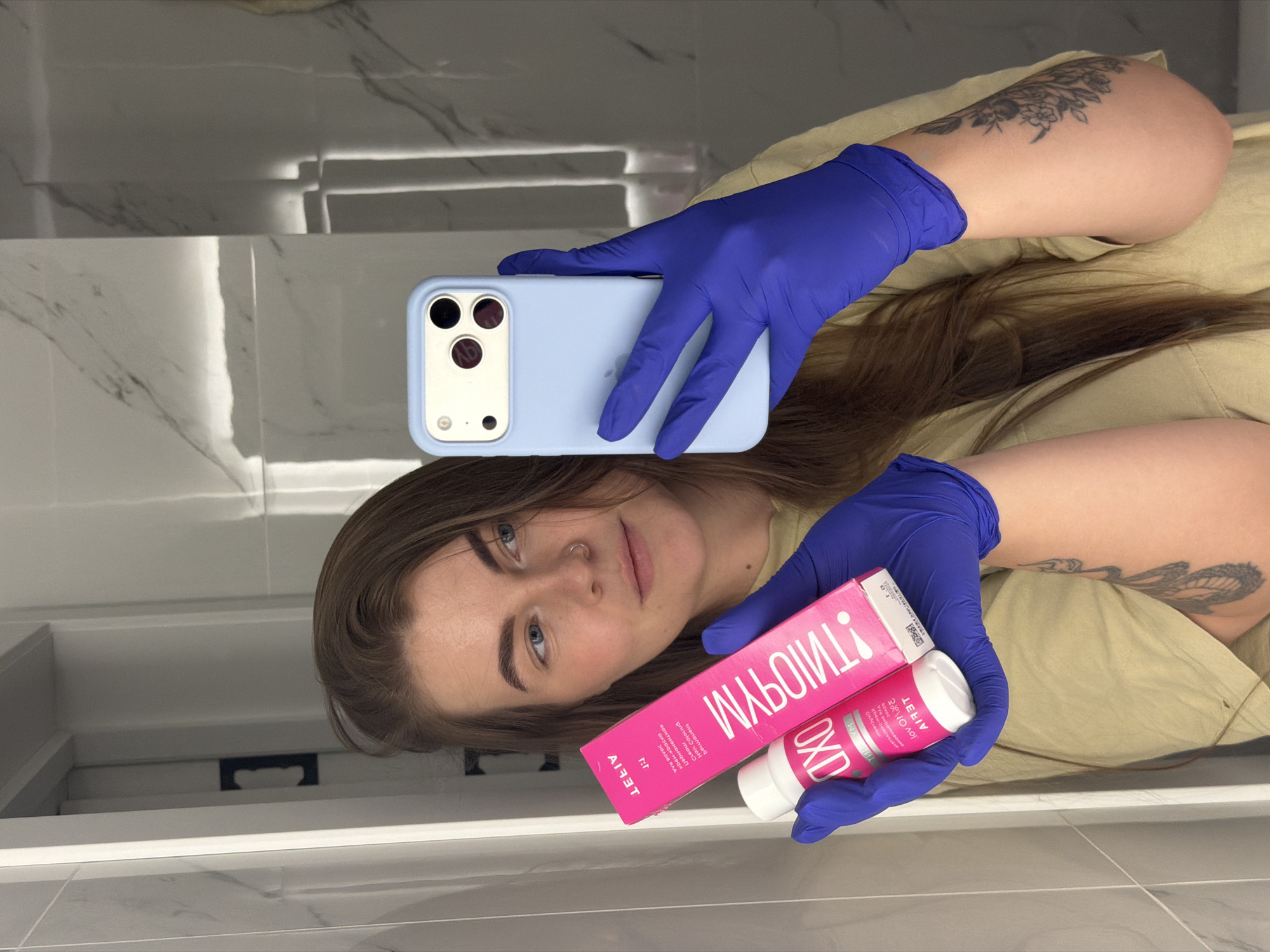 The image shows a person, possibly female, holding a mobile phone and a cosmetic product in gloved hands, against the backdrop of a bathroom setting. The person has fair skin, light eyes, and brown hair. Tattoos are visible on both arms. The phone is encased in a light blue case. The cosmetic product, consisting of a box and a bottle, features pink packaging with lettering that appears to be partially mirrored. The gloves are a bright shade of blue. The bathroom environment includes white and grey marble-like tiles, a white sink or countertop, and what appears to be a white drawer or shelf beneath. The location is specified as the city of Chaikovsky, Russia.