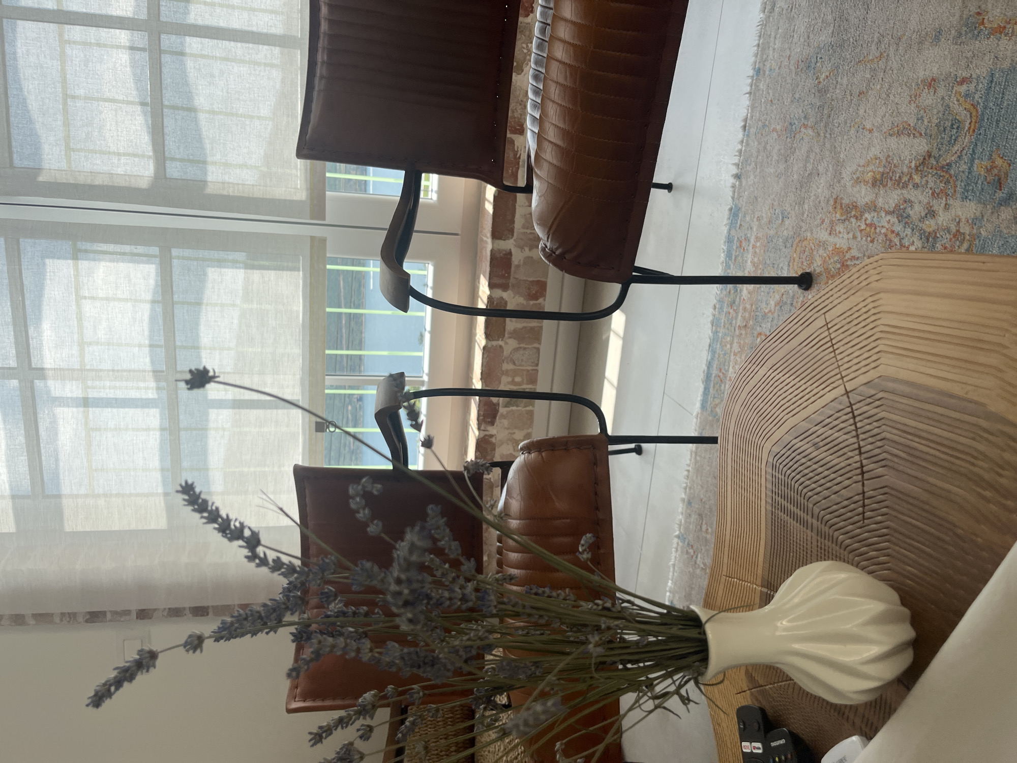 The image depicts a still life arrangement in a modern interior, likely a living room in Kumbor, Montenegro. In the foreground, a bouquet of dried lavender in a textured white vase sits on a light-colored, slatted coffee table. The lavender stems are long and delicate, with visible dried florets.
Behind the coffee table, two brown leather chairs with a distressed, vintage appearance are positioned near a large window covered with sheer white curtains. The chairs have dark metal frames and armrests. Through the window, a glimpse of the outdoors can be seen, suggesting a bright, daytime scene, possibly with fair weather. The light filtering through the curtains creates a soft, diffused illumination.
The flooring appears to be a light wood or laminate. A portion of an area rug with a muted, abstract pattern in shades of blue, beige, and orange is visible between the chairs and the coffee table. The backdrop shows exposed brickwork behind the chairs, adding an element of rustic charm to the otherwise contemporary setting. The overall mood is serene and inviting, with a focus on natural materials and textures. No people are visible in the image. There is some visible text on a remote control device on the table.