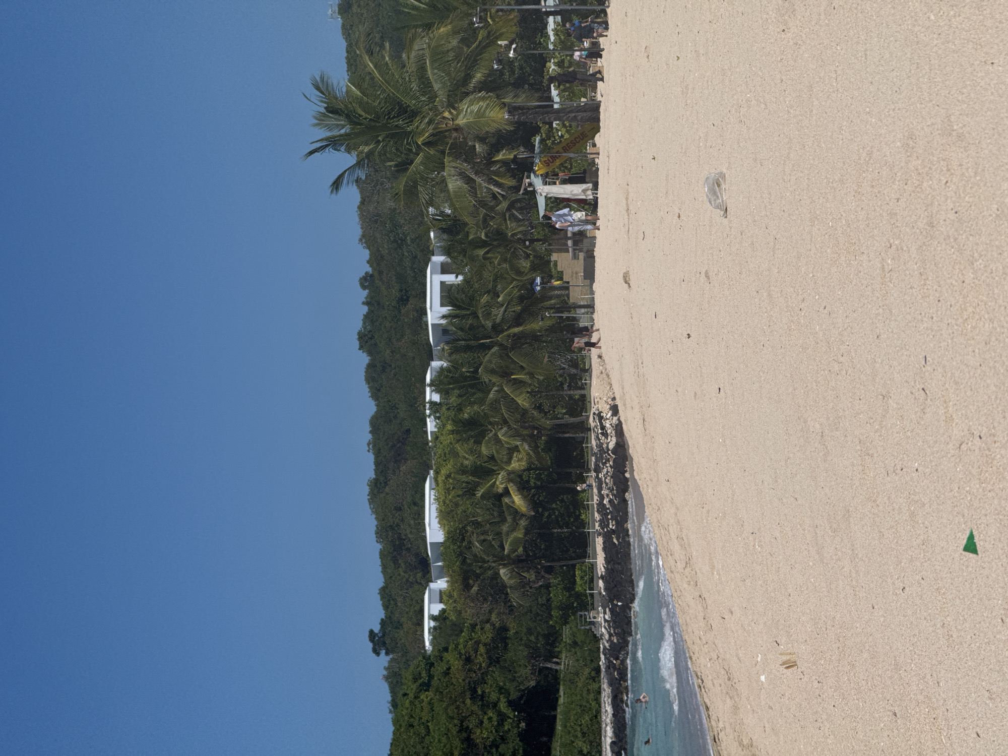 The image shows a beach on a sunny day. The sky is clear blue. The beach is sandy, with some debris scattered around. A line of trees and vegetation separates the beach from a set of white buildings on a small hill. People can be seen in the distance along the beach. The location of the image is in an Unknown city.