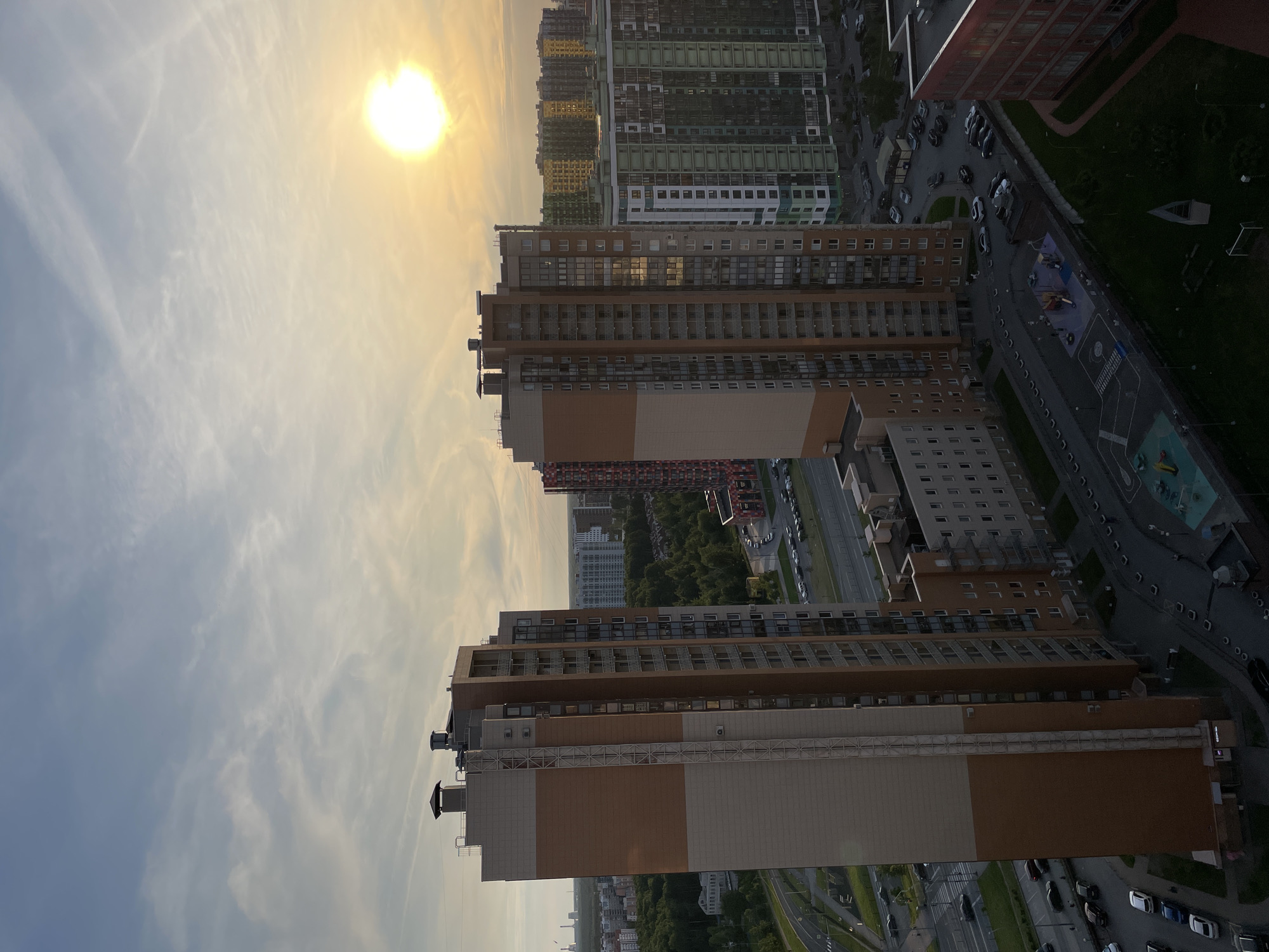 The image depicts an aerial view of a residential area in the outskirts of a city, likely during late afternoon or early evening, indicated by the low sun. The foreground is dominated by two tall, modern residential buildings. The building on the left is primarily light beige with prominent vertical bands of orange-brown. The building on the right is a similar height and design, predominantly beige with more subtle vertical detailing and numerous balconies.
Behind these buildings, a complex of other multi-story residential buildings is visible, showcasing a variety of colors including green, yellow, and red. These structures are densely packed, suggesting a large housing development. A road network with cars is visible in the mid-ground, winding between the buildings and leading towards a treeline in the distance. There are also indications of infrastructure like power lines and chimneys.
In the lower part of the image, a playground area with colorful equipment is visible, along with pathways and parked cars. The sky is a mix of blue and pale orange, with scattered clouds, and the sun is a bright, diffused orb in the upper right quadrant. The overall impression is one of urban expansion into a formerly more natural landscape, with a focus on high-density housing.