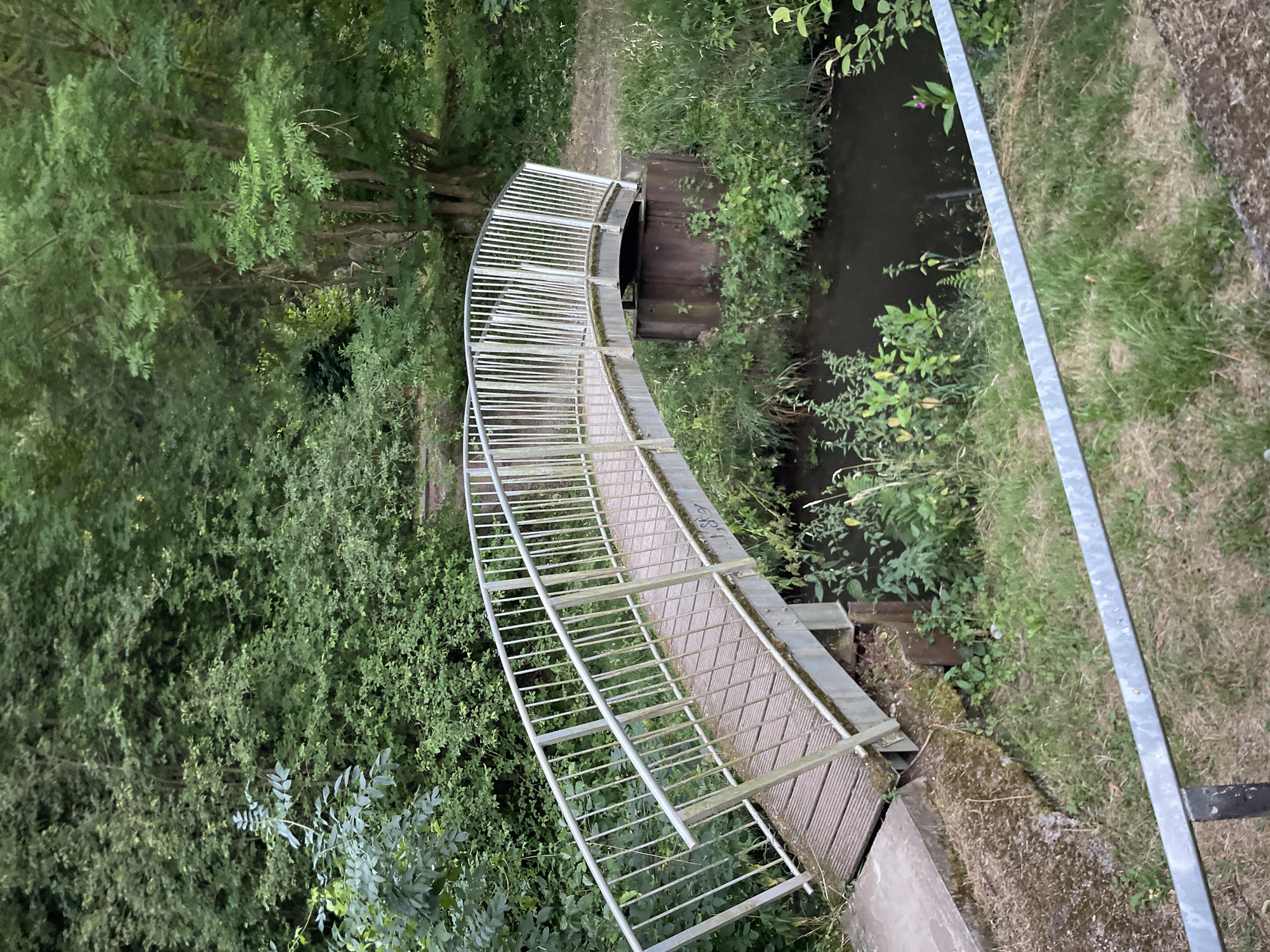 A slightly overcast, medium shot captures a curved, white-railed bridge constructed with wooden slats that span a narrow, dark-water stream. The bridge, likely made of metal, is supported by metal beams and rests on a rusty metal cylindrical foundation on the right side and a concrete structure on the left. The stream bank is densely populated with lush green foliage and grasses. Behind the bridge, a dense forest of trees with varying shades of green foliage forms a natural backdrop. The path leading to and from the bridge is visible, appearing as a dirt trail. The scene is devoid of human presence or any discernible activity. The overall ambiance suggests a peaceful, natural setting within a park or wooded area. The location is identified as Neukirchen, Germany.