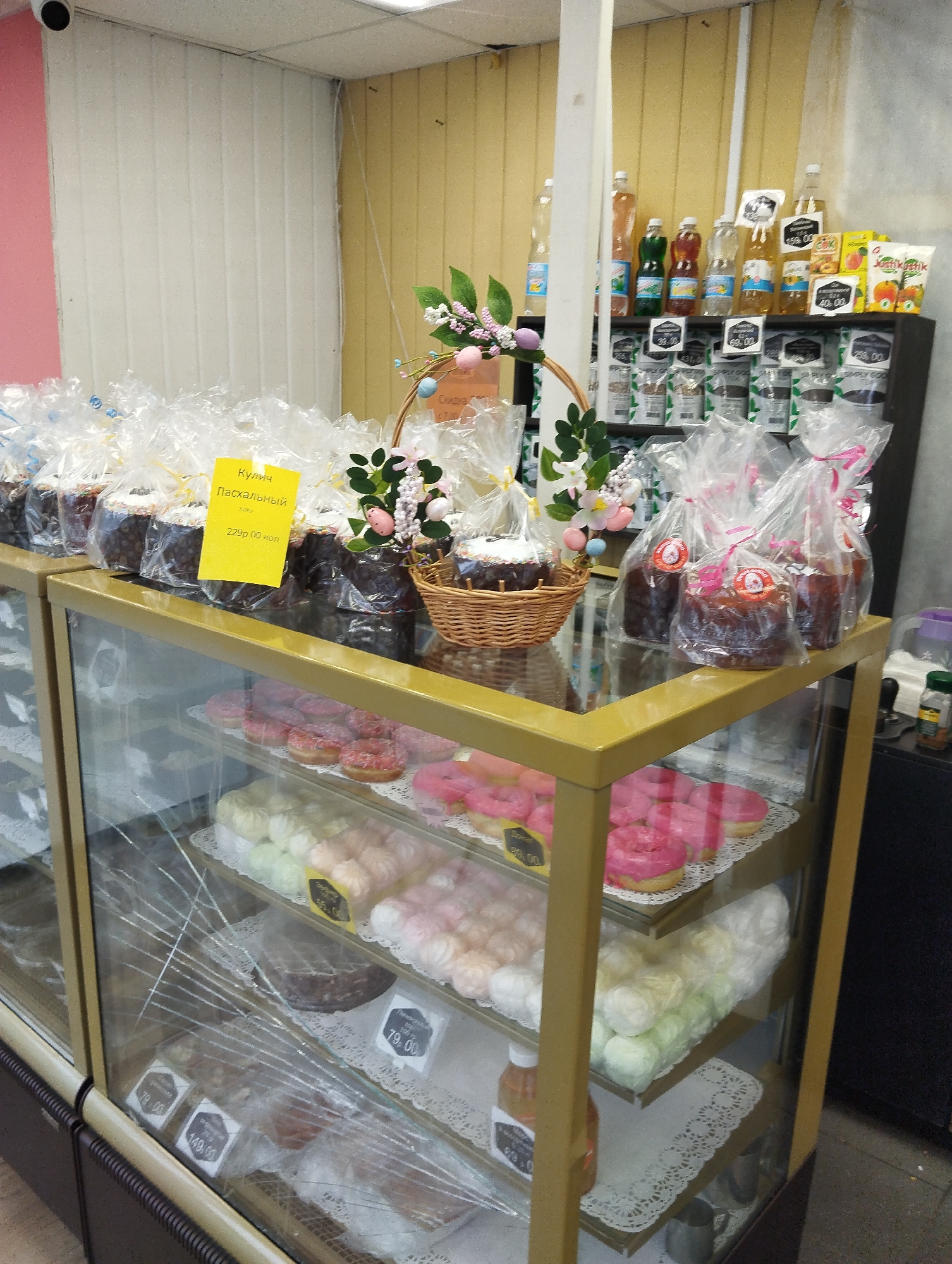 The image depicts a retail setting, likely a bakery or grocery store, in what is possibly Votkinsk, Russia, based on the Cyrillic text visible. A glass display case dominates the foreground, showcasing various baked goods. On the top shelf, wrapped Easter cakes are arranged, with a yellow price tag displaying "229p 00 коп" (229 rubles, 00 kopeks). A decorative Easter basket containing similar cakes is placed centrally on the top. Below, the shelves feature doughnuts of varying colors and marshmallows. The glass of the display case appears to be cracked. Further to the back, on a higher shelf, are more baked goods and jars, potentially other products for sale. A black shelf unit in the background holds a variety of bottled beverages, possibly juices and teas. The environment appears to be a typical small retail space with basic lighting and a neutral color scheme. The absence of people in the frame suggests the image was taken either before or after business hours, or in an uncrowded moment.