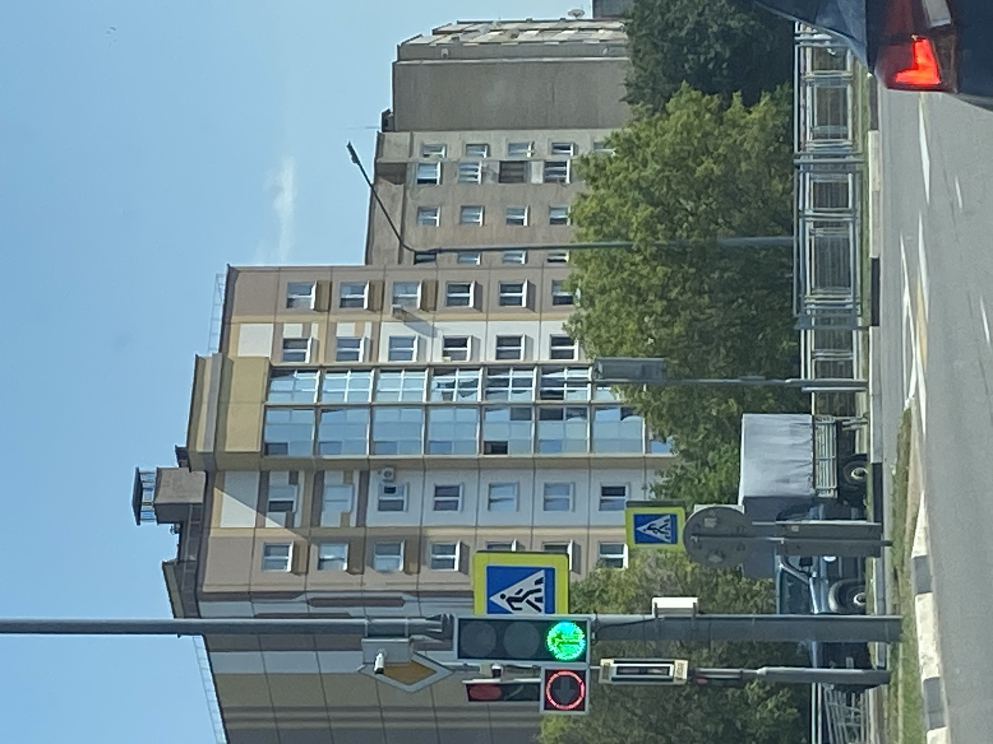 This is a street-level, eye-level, medium shot photograph of a traffic intersection in Brest, Belarus. The foreground shows a street with white pedestrian crossing lines. To the right, the rear taillight of a dark-colored car is visible. A traffic light pole with red and green signals, and a pedestrian crossing sign are prominently displayed on the left side of the image. A dark-colored pickup truck with a white tarp-covered cargo bed is stopped at the intersection. In the background, multi-story apartment buildings of varying architectural styles and colors are visible, along with green trees. The sky is clear and blue.