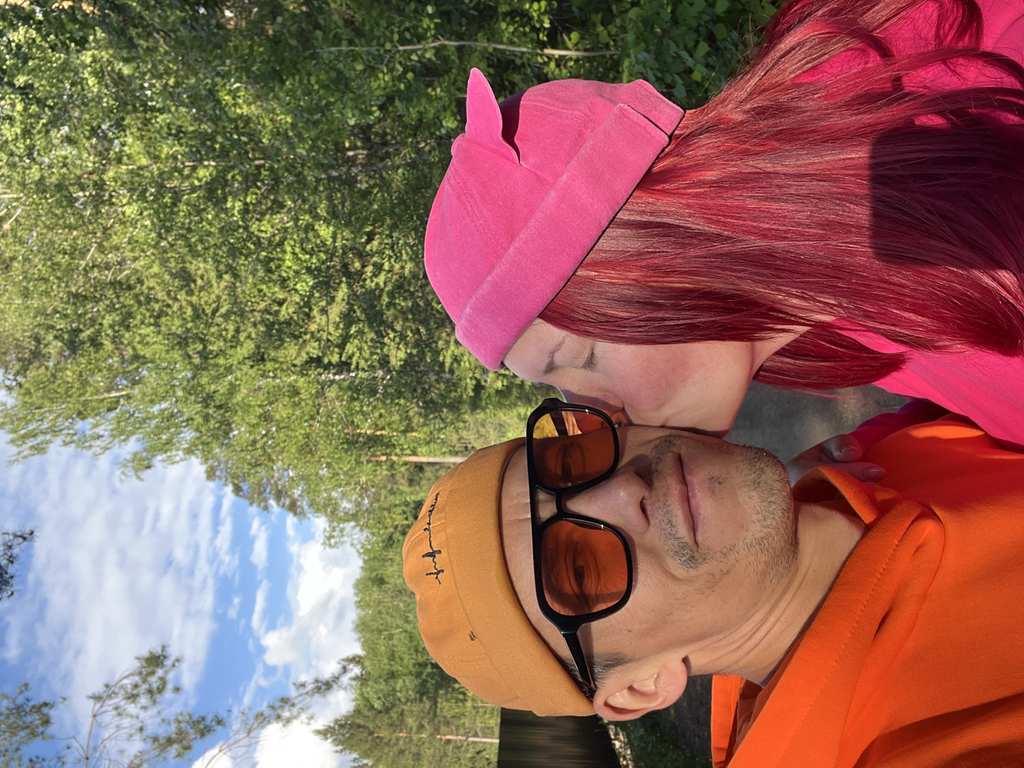 The image, originally rotated 90 degrees clockwise, captures a close-up, affectionate moment between two individuals outdoors in Chaikovsky, Russia. The scene depicts a man and a woman in a forest or park setting under a clear blue sky with scattered white clouds.
The man, positioned on the left (when viewed upright), wears an orange hoodie and an orange or light brown beanie with a small, indistinct dark embroidered design. He has short stubble and is wearing black-framed sunglasses with orange-tinted lenses that reflect the surrounding trees and sky. His expression is neutral as he looks forward.
The woman, on the right, has long, vibrant red or magenta hair, part of which is covered by a bright pink beanie. She is wearing a pink top or hoodie and is leaning in to kiss the man on his cheek, with her eyes closed, suggesting a tender moment. Her hand is visible, gently resting on his shoulder or chest.
The background consists of dense green foliage and trees, indicating a natural, wooded environment. The bright, natural lighting suggests it is daytime, likely a sunny morning or afternoon. The overall mood conveyed is one of affection and connection in a serene outdoor setting. There is no clearly discernible text visible on the subjects' attire.