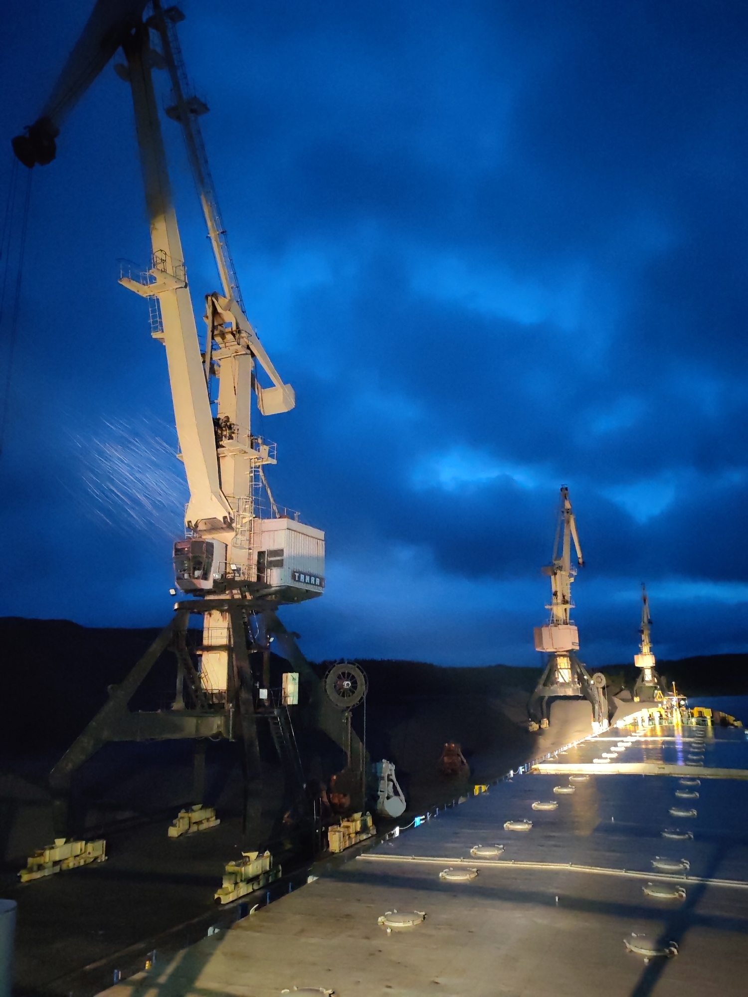 The image depicts a port scene at night. Three large cranes are visible, the largest being in the foreground with water spraying from its arm. The cranes appear to be at the port of Shalsky, Russia. The deck of a ship is visible with several hatches, implying the scene is taken from the ship’s deck. The sky is dark blue with clouds. A dark landmass, possibly a hill, is in the background.
