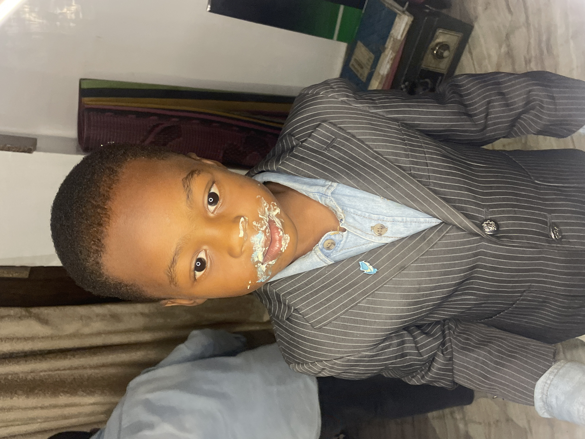 A young child with dark skin and short hair is wearing a pinstripe suit over a light blue denim shirt. He has frosting around his mouth, indicating he may have been eating cake. The background includes furniture and objects such as books and a small safe or strongbox. The location, Jos, Nigeria, cannot be determined from the visual details of the image.