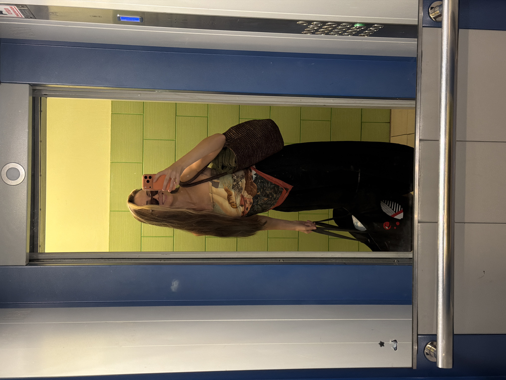 A young woman is taking a mirror selfie in an elevator. She is wearing sunglasses, a patterned scarf as a top, and black pants. She is holding an orange smartphone in her right hand and a brown woven bag on her right shoulder. In her left hand, she is holding a black tote bag. The elevator has blue and white walls, and the interior of the elevator car is visible in the mirror, featuring green tiled walls and a yellow ceiling. The elevator appears to be located in Khimki, Russia.