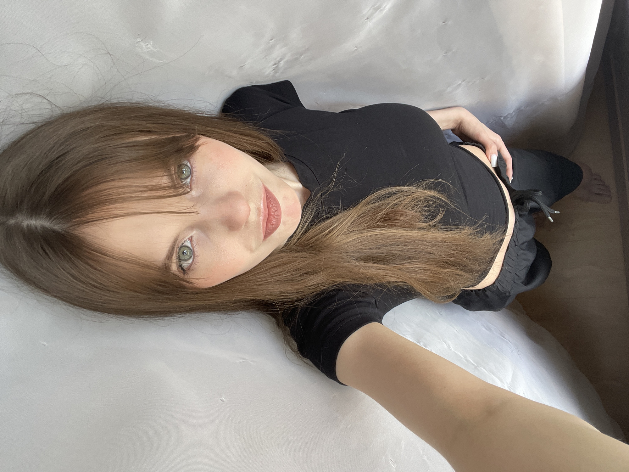 A young woman with long brown hair and bangs, wearing a black top and pants, is lying down on a white surface. She is looking up at the camera with a slight smile. The setting appears to be indoors, possibly on a bed or soft surface. There is no verifiable evidence in the image to indicate the location is Ufa, Russia. The surrounding environment is nondescript.
