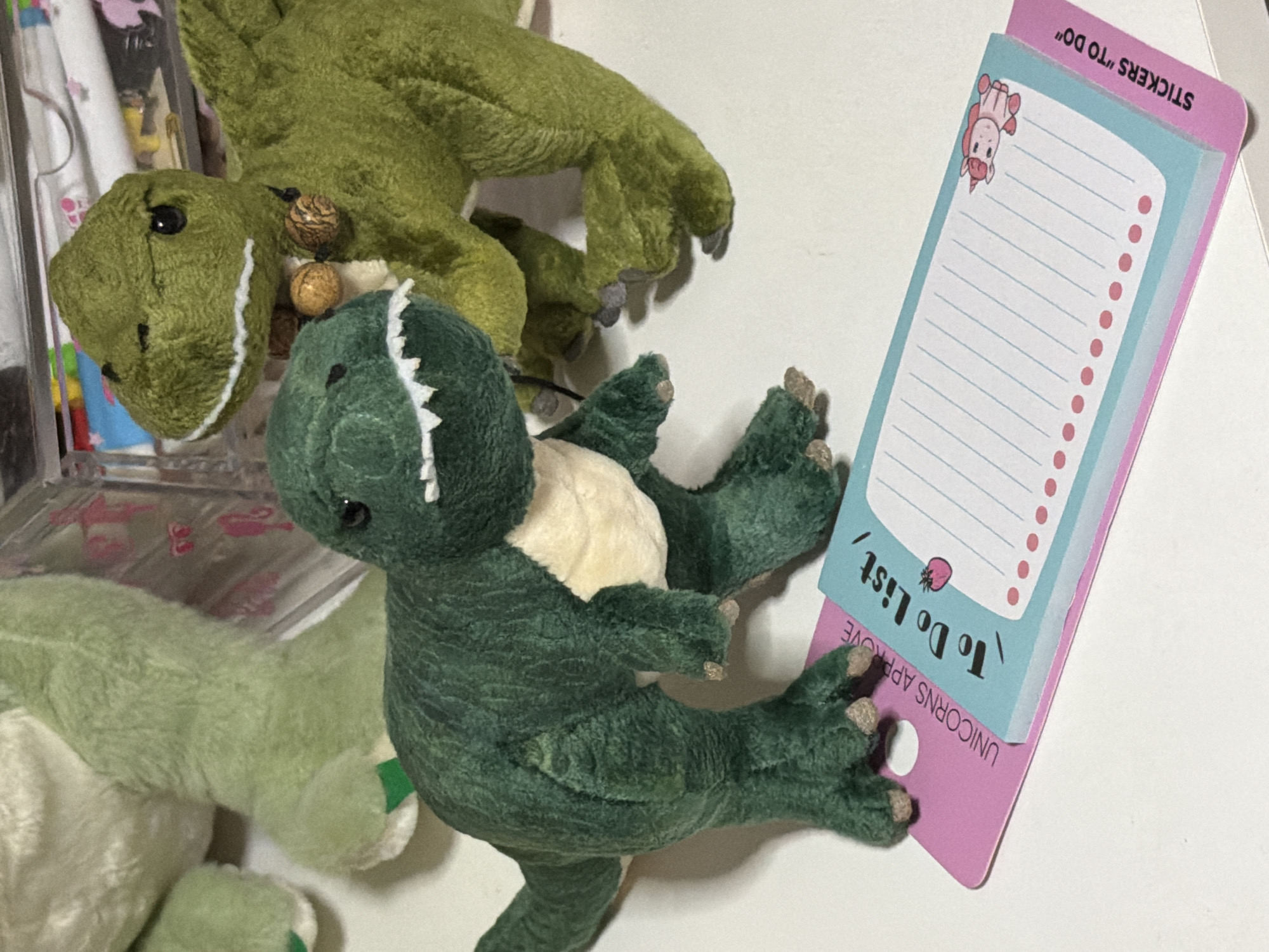 The image shows a close-up, eye-level, indoor shot of several green plush dinosaur toys and a pink and blue notepad on a white surface. In the foreground, a dark green plush dinosaur is facing forward with its mouth open, revealing white teeth. Behind it and slightly to the right, a lighter green plush dinosaur is also facing forward, wearing a collar with two small, round brown beads. To the left of the dark green dinosaur, a portion of another light green plush dinosaur is visible. A pink notepad with "To Do List" written in stylized black font is placed in front of the dark green dinosaur. The notepad has blue lined paper, pink dots along the bottom, a small strawberry illustration, and a cartoon unicorn in the bottom right corner. The phrase "UNICORNS APPROVE" is printed vertically on the pink backing of the notepad, and "STICKERS 'TO DO'" is printed vertically on the bottom right edge of the pink backing. The background consists of a blurred clear container with various colorful items inside, possibly art supplies or stationery. There is no indication of the location being in Moscow, Russia.