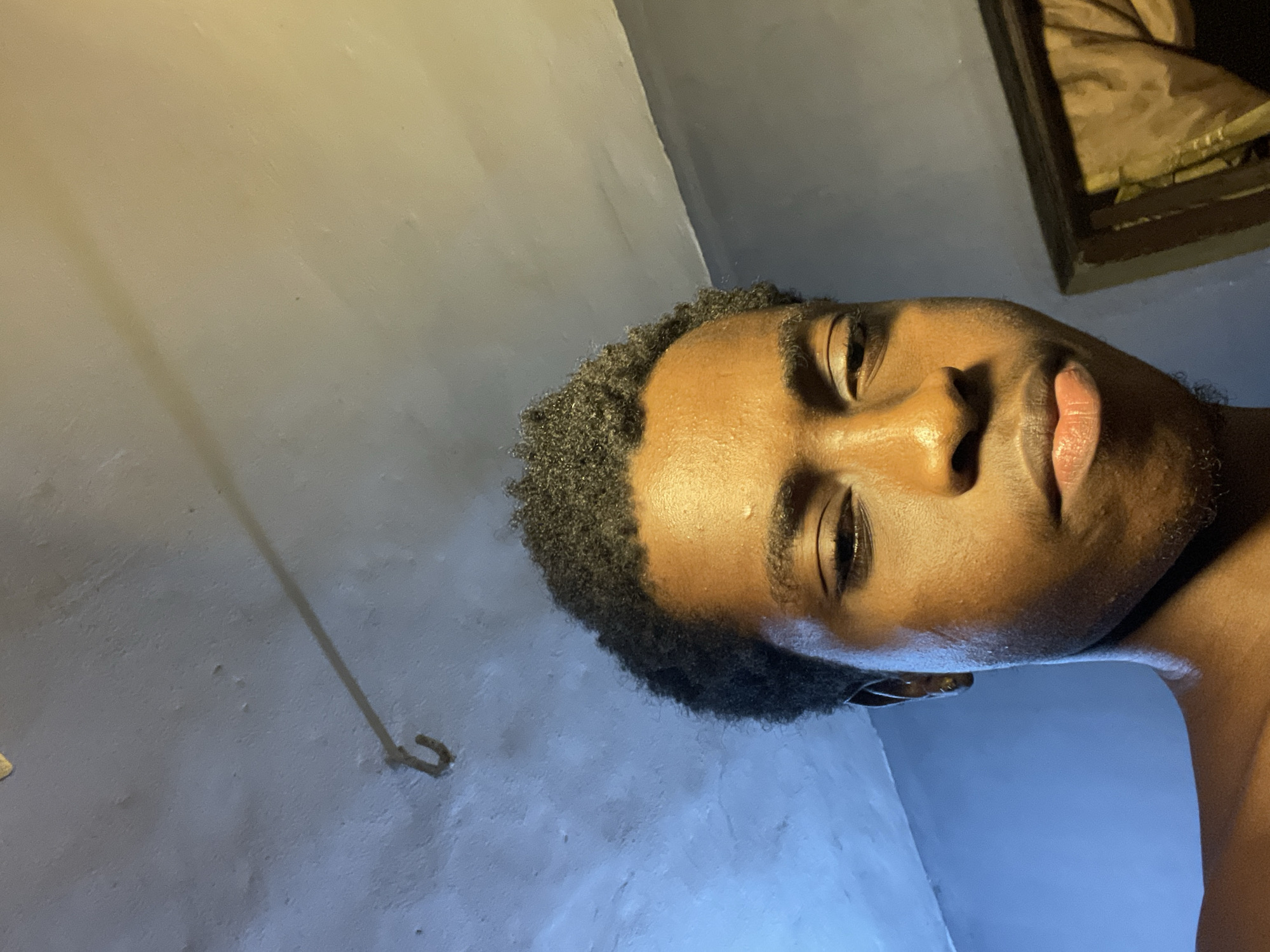 A low-angle, rotated close-up shot of a dark-skinned man indoors. His head is mostly visible, with a portion of his shoulder appearing in the lower right. He has short, dark, curly hair. His facial features are prominent: eyes, nose, lips. A small earring is visible in his left ear. The background consists of a pale, unevenly lit wall. A metal hook is visible towards the left. In the upper right corner, there is the edge of a framed object. The location is indiscernible; there are no elements within the image that allow for a specific identification of the city of Nkpor, Nigeria.