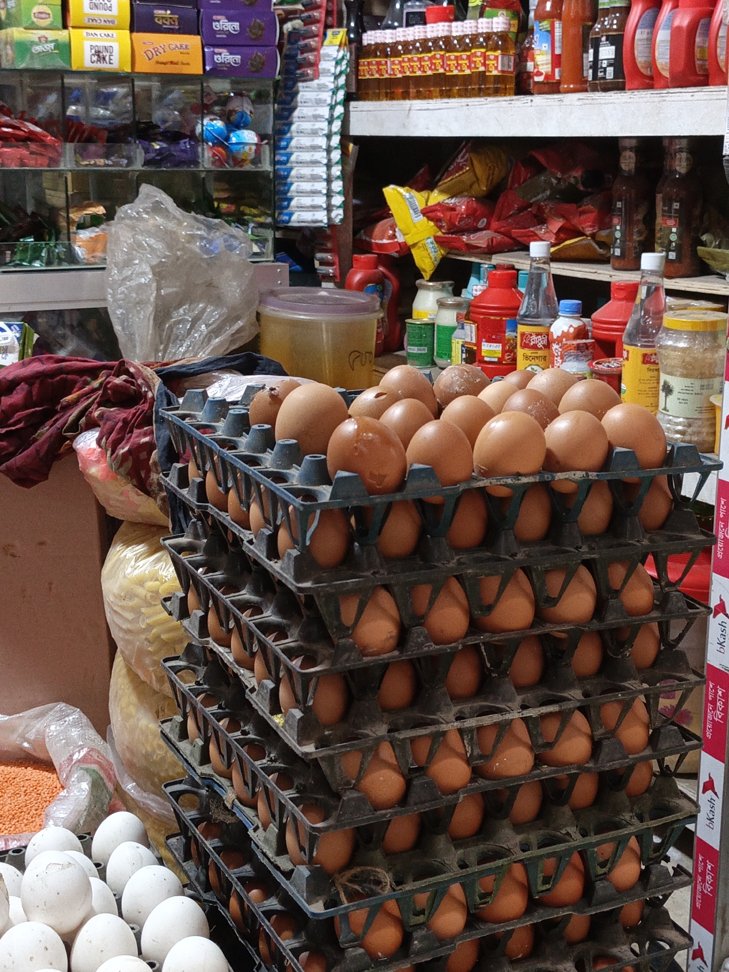 This vertical shot captures a busy display of goods, primarily focusing on a large stack of eggs in a market or grocery store setting. In the foreground, numerous brown eggs are meticulously arranged in multi-tiered black and blue plastic trays, stacked at least seven levels high. To the bottom left, a pile of white eggs (likely duck eggs) is also visible. Next to the white eggs, a loose pile of orange lentils or dal can be seen.
The background reveals shelves laden with various packaged food items. On the upper shelves, bottles of sauces and condiments, many with red caps and labels, are neatly lined up. Below them, a shelf holds numerous bags of snacks and chips, some red and yellow, appearing somewhat dishevelled. Higher up, packaged cakes and biscuits in yellow and purple boxes are visible. A transparent plastic bag sits on a counter in the middle ground, near some folded maroon fabric.
Visible text on the product packaging includes "POUND CAKE," "DAN CAKE," and "DRY CAKE" on yellow boxes, and "অজন" (Ojon) and "ওরিনো" (Orino) on purple boxes. Bottles display "রাঁধুনী ভিনেগার" (Radhuni Vinegar). On the right side of the image, red and white promotional strips bear the Bengali text "প্রয়োজনে পাশে" (Proyojone Pashe - "By your side when needed") and "বিকাশ" (bKash), along with the English "bKash," indicating the presence of a mobile financial service often seen in Bangladesh. The lighting suggests an indoor environment, likely during the day, with no people visible.