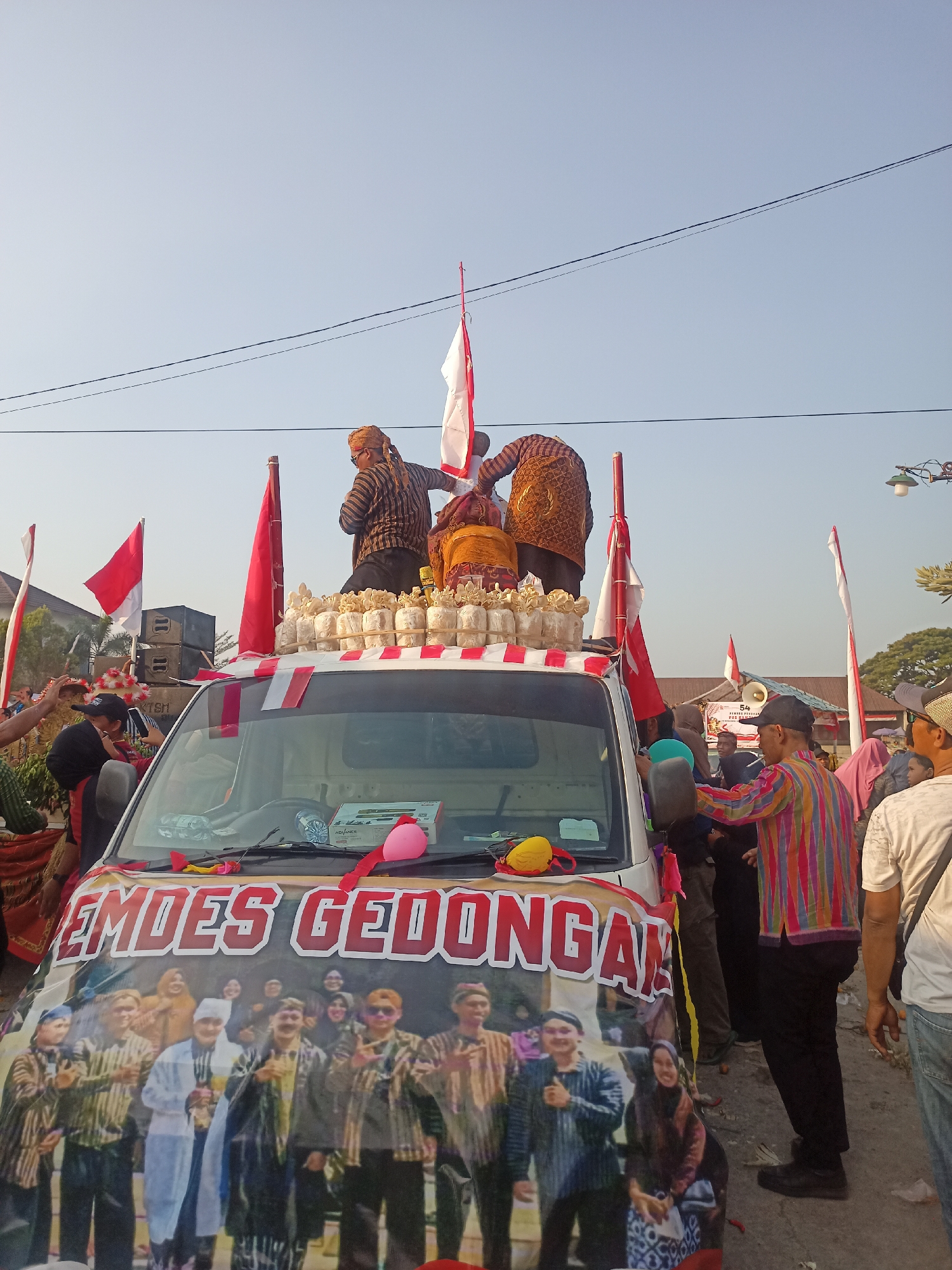 This is a medium shot, eye-level, outdoor, daylight, full-color, slightly over-saturated photograph taken on a street in Ngrompongan, Indonesia. A white pickup truck is the central subject, decorated with a large banner on its hood featuring the words "EMOES GEDONGAN" and a montage of people. Several individuals are standing on the flatbed of the truck, some wearing traditional Indonesian attire, including batik shirts and head coverings. They appear to be part of a parade or celebration, holding red and white Indonesian flags. The truck is adorned with red and white striped tape and other decorative elements. A crowd of people is visible on the ground surrounding the truck, some interacting with those on the truck. In the background, there are buildings, trees, and power lines, indicative of a populated urban or semi-urban environment. The sky is clear and blue, suggesting fair weather. The overall scene conveys a festive and communal atmosphere.