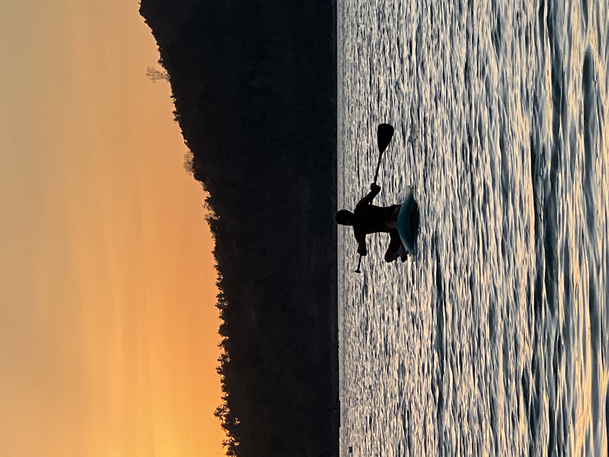The image depicts a serene scene of a person stand-up paddleboarding on the water at sunset. The silhouette of the paddleboarder, holding a paddle, is clearly visible against the rippling water. The water reflects the warm hues of the sunset, creating a textured and dynamic surface.
To the left of the frame, the dark silhouette of a tree-lined hillside rises against a soft, gradient sky transitioning from pale orange to a lighter hue at the top, indicating dusk. The overall mood is peaceful and tranquil, with the natural beauty of the sunset and the calm water creating a sense of relaxation.
The location is identified as Ko Pha-ngan District, Thailand, which aligns with the tropical imagery of lush vegetation on the hillside and the warm, inviting sunset. No visible text is present in the image. The time of day is clearly sunset, and the weather appears to be clear and calm, ideal for paddleboarding. The activity is recreational, enjoying the outdoors at the end of the day.