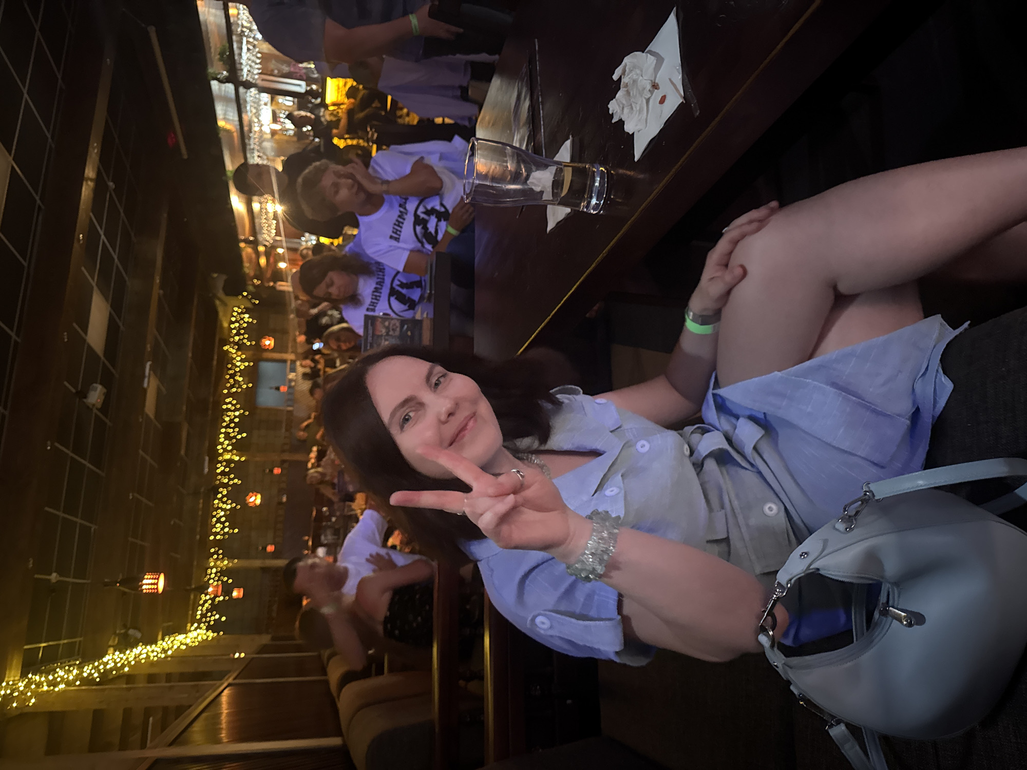 Here's a description of the image:
**General:** The image shows a woman posing for a photo inside what appears to be a restaurant or bar.
**People:**
* The woman in the foreground is making a peace sign. She has dark hair and is wearing a light blue button-down shirt and a bracelet.
* In the background, there are numerous people seated at tables. They appear to be socializing. Some are wearing t-shirts with writing.
**Objects:**
* A table is visible, with a glass and some napkins on it.
* A light blue purse is near the woman.
* String lights are visible in the background.
* There are bar stools in the background.
**Actions:**
* The woman is posing for a photo.
* People in the background are socializing, sitting at tables.
**Environment:**
* The location is a restaurant or bar, based on the tables, bar stools, and atmosphere.
* There is string lighting, creating a warm ambiance.
* There is what appears to be an empty bar or countertop behind the people in the background.
**Location:**
* There is no visible information in the image that can be used to determine that this photo was taken in Chaikovsky, Russia.