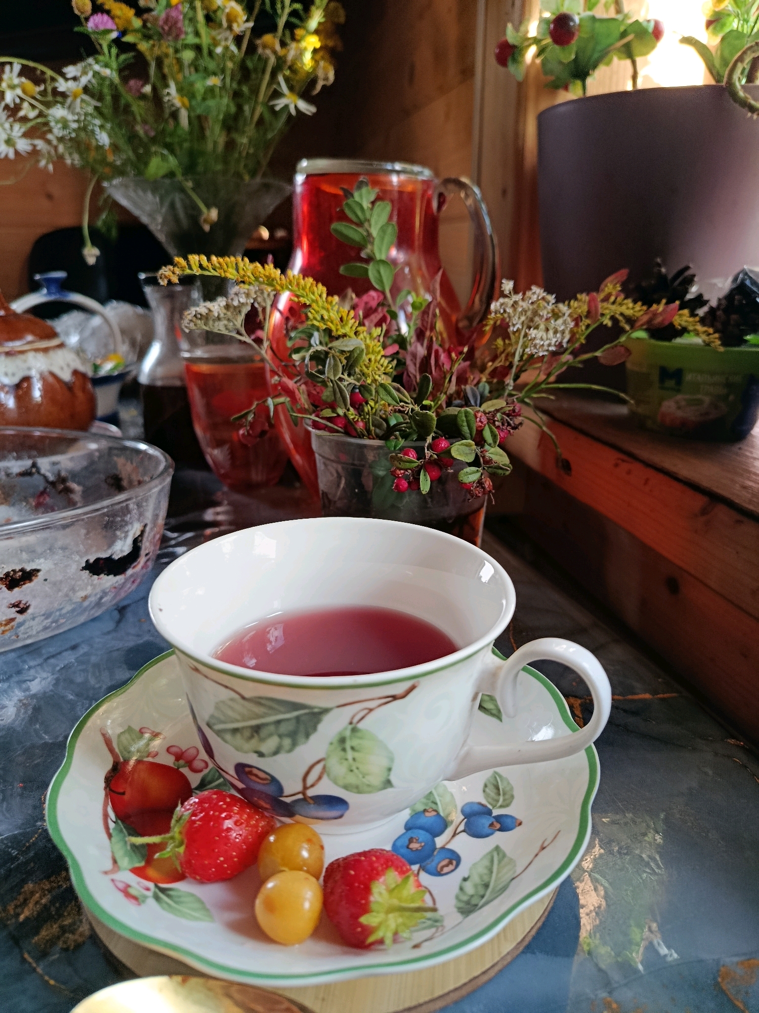 A close-up shot shows a white teacup filled with a red liquid, placed on a saucer. The saucer is decorated with illustrations of fruits, including strawberries, blueberries, and yellow berries, as well as green leaves. Beside the teacup and saucer, on the left, is a clear glass bowl containing remnants of what appears to be dark-colored food. In the background, a pitcher of red liquid is visible, along with a vase of wildflowers. Further in the background, there is a wooden surface and a dark-colored pot. The scene is set in an indoor environment, possibly a room with wooden walls, with natural light illuminating the objects. The overall ambiance suggests a relaxed setting.